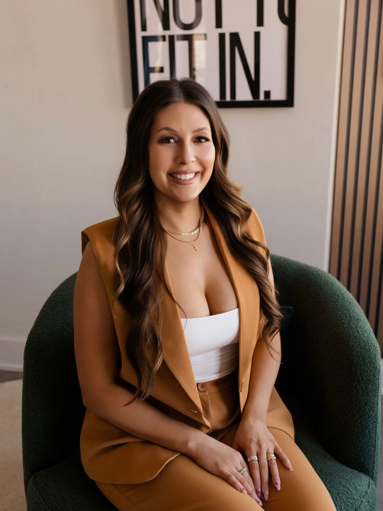 A woman sitting on a dark green chair in a room with a white wall and a framed sign behind her that reads 'NOT TO ETT IN.' She has long, wavy brown hair, is smiling, and is wearing a tan sleeveless blazer, white tank top, matching tan pants, layered necklaces, and rings on her fingers.