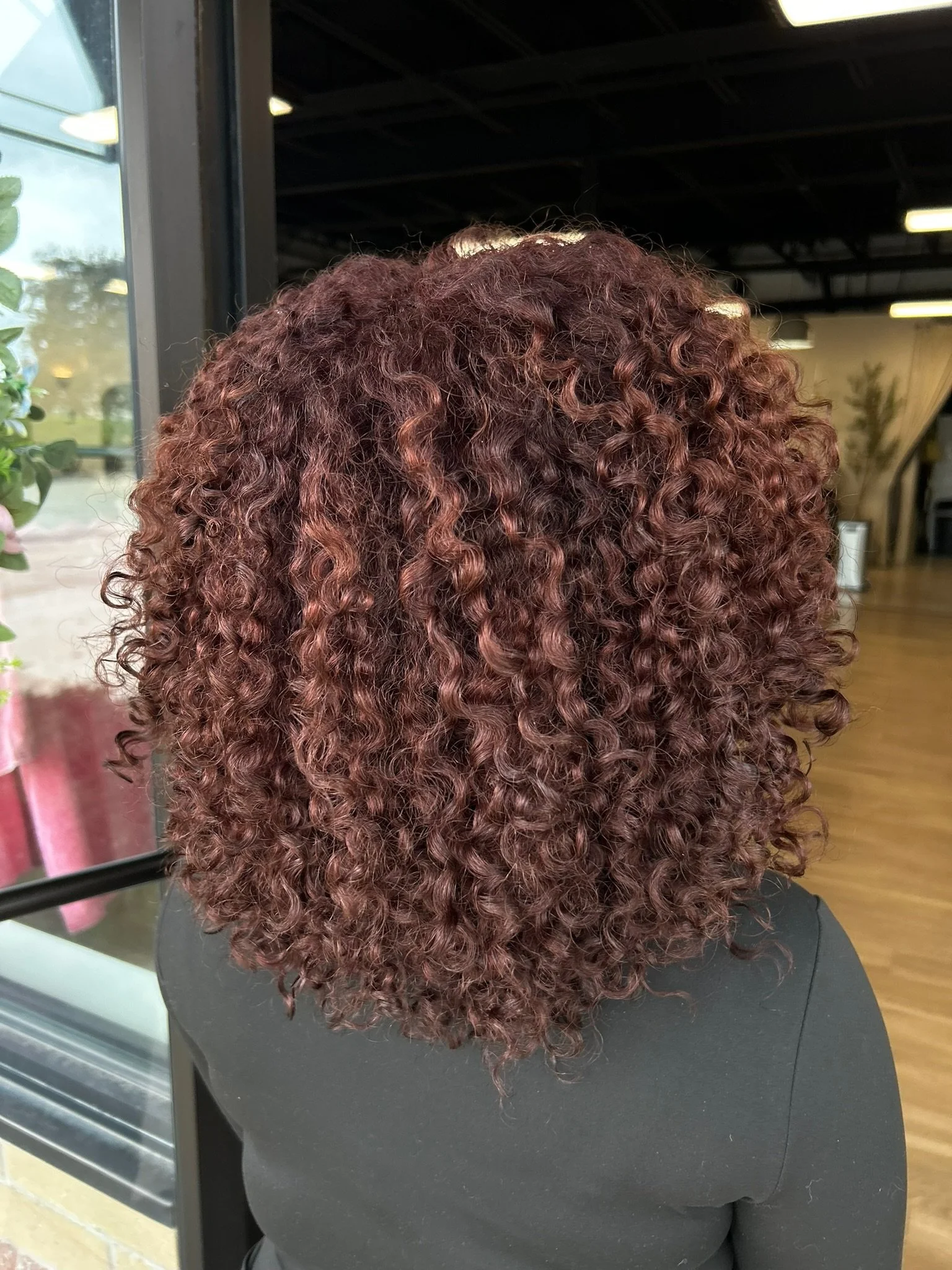 Reddish-brown curls viewed from behind over a black top near the salon window.