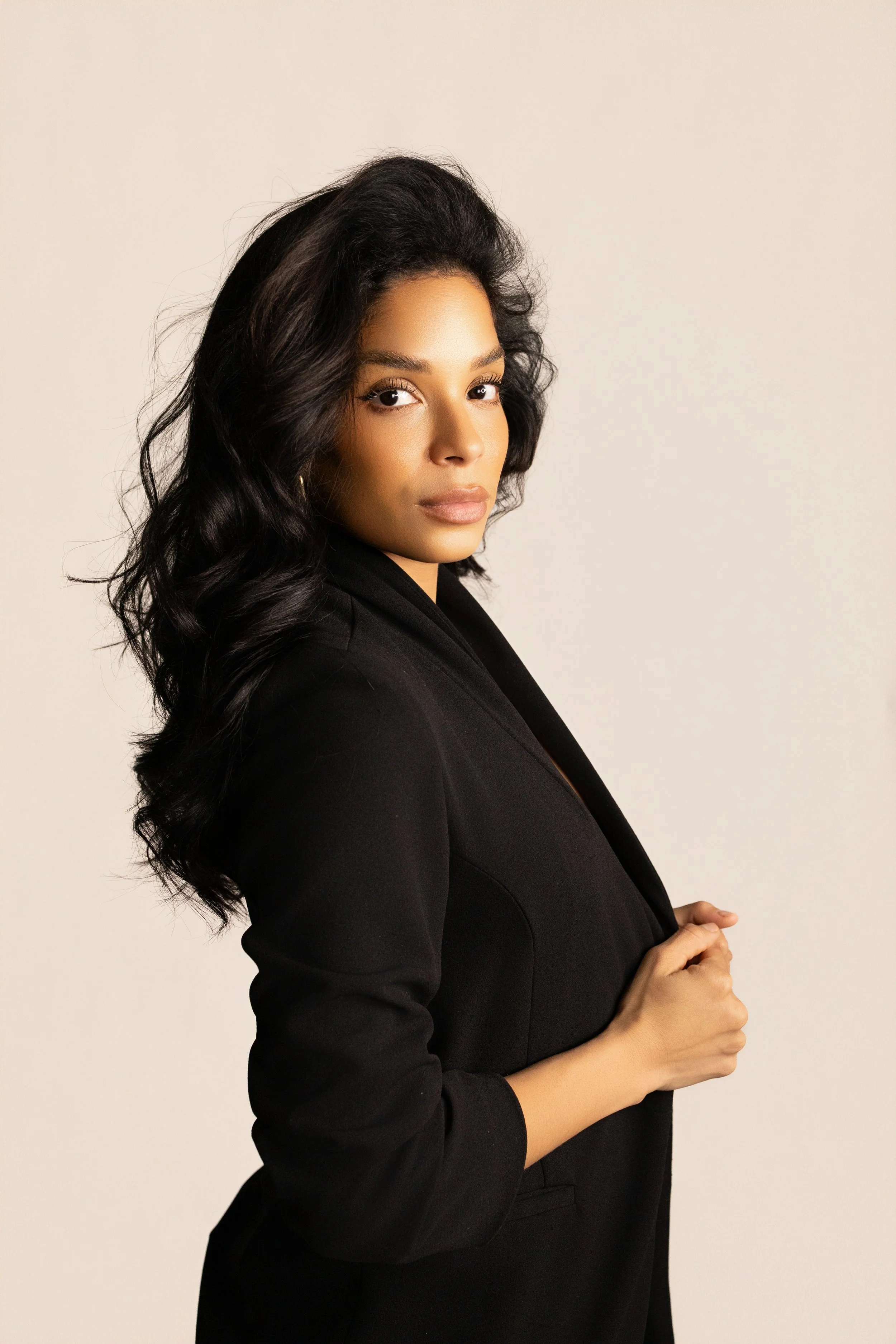 A confident woman with braided hair wearing a white suit jacket, matching white pants, a black crop top, and layered gold necklaces stands against a plain white background.
