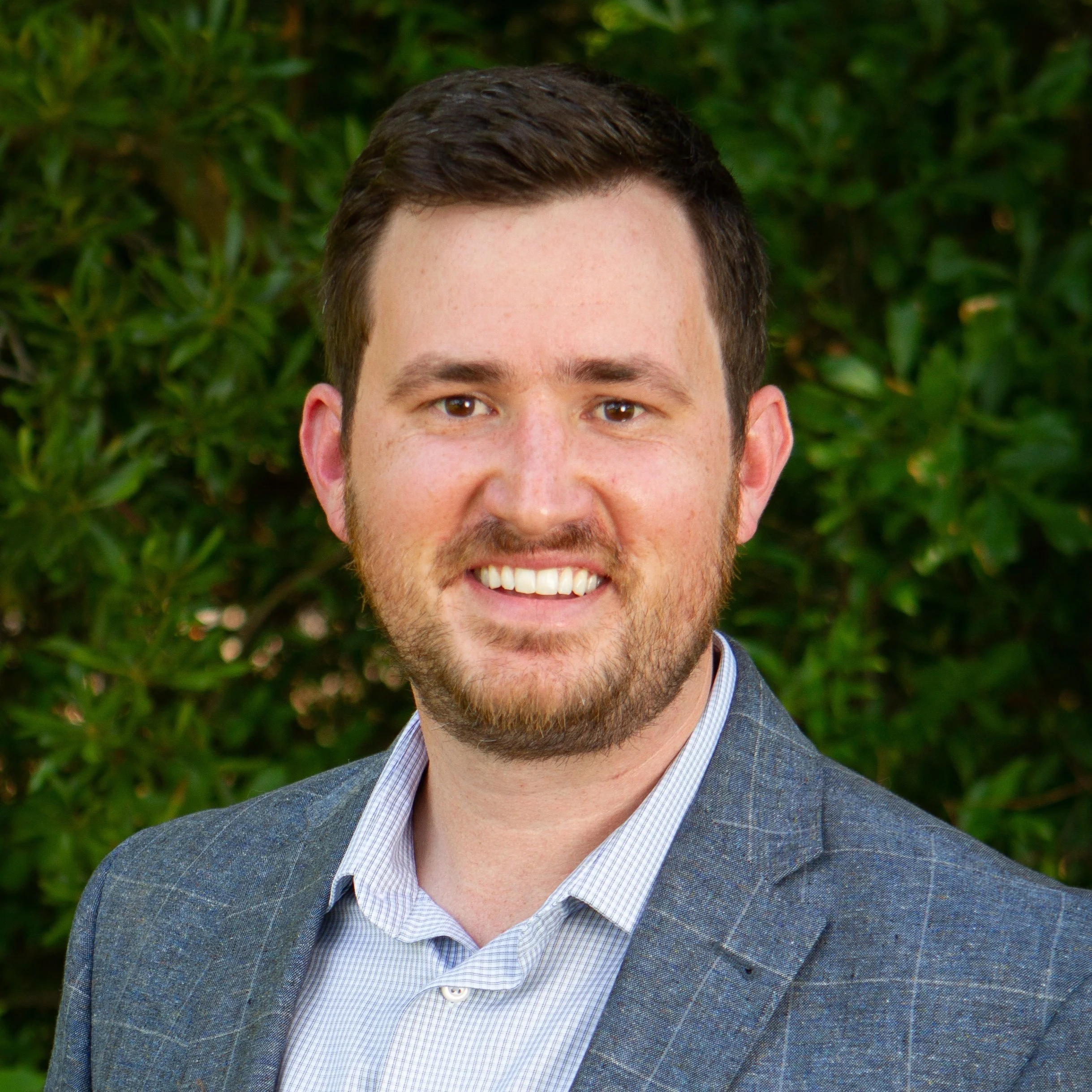 Headshot of a young man with dark brown hair, light beard, smiling, wearing a gray plaid blazer over a light collared shirt, outdoors with green foliage in the background.