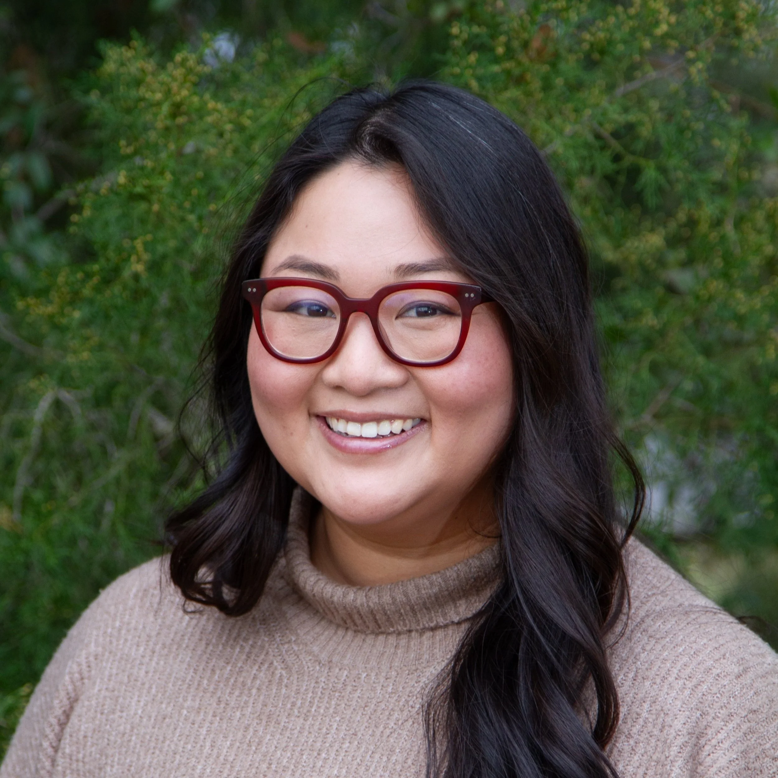 A smiling woman with long dark hair, red glasses, and a beige turtleneck sweater outdoors with green trees in the background.