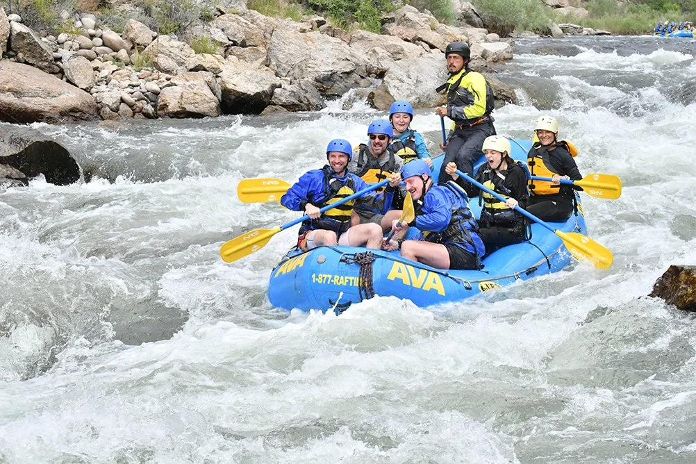 Group of people whitewater rafting in a blue inflatable raft, wearing helmets and life jackets, navigating through rapids.