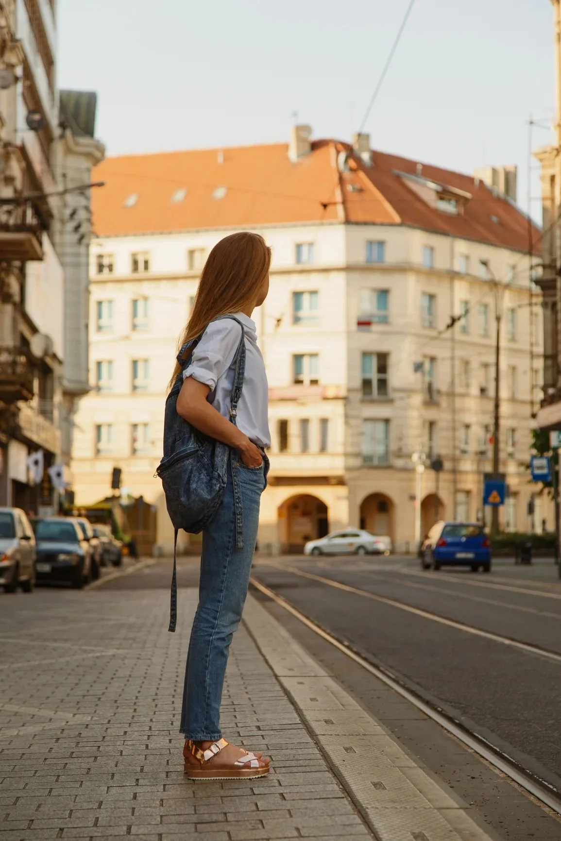 A woman wearing jeans, a button-down shirt, platform sandals, and a backpack is standing on a Prague street.