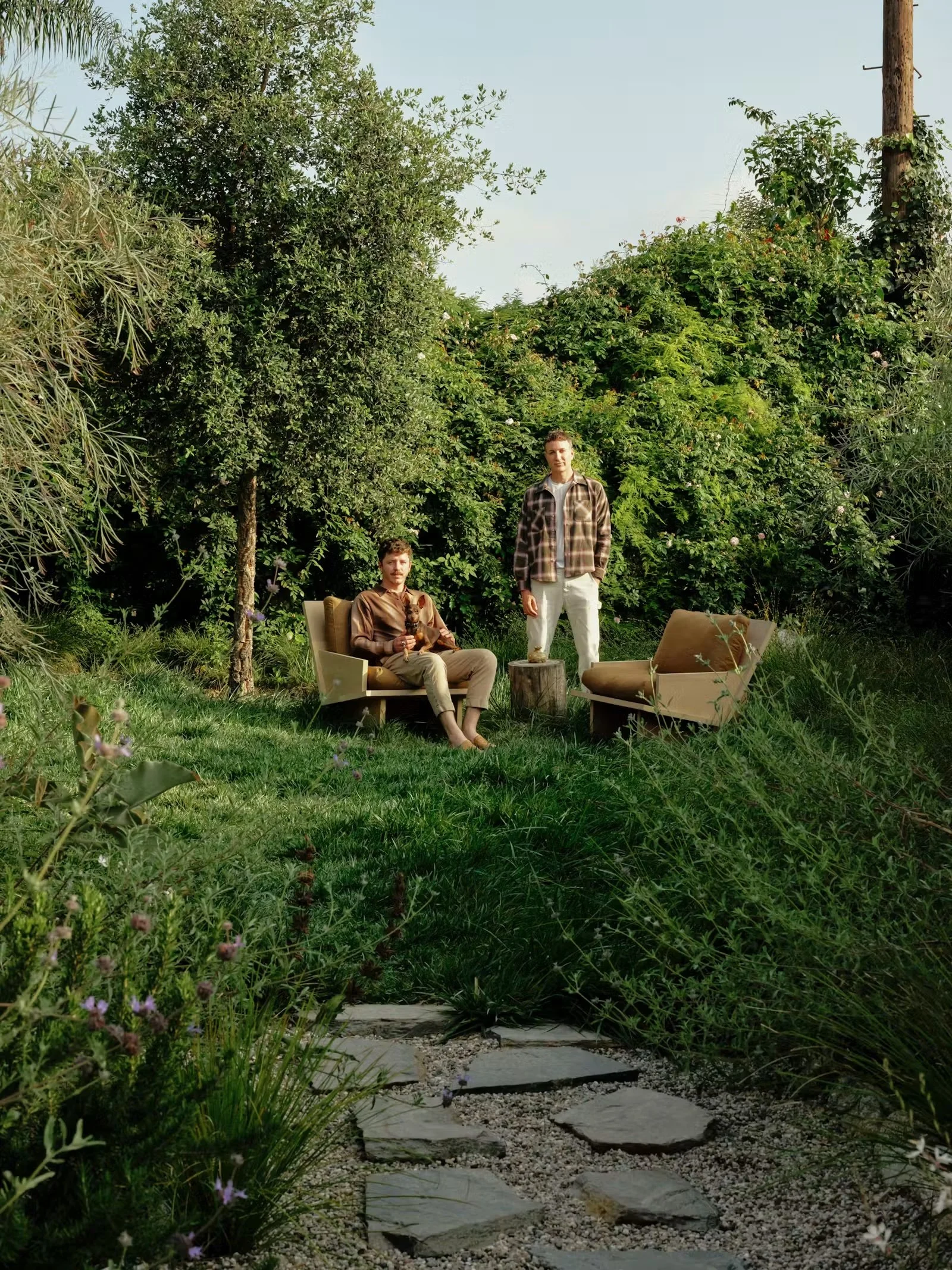 Two men are standing (one is sitting) in their lush backyard.