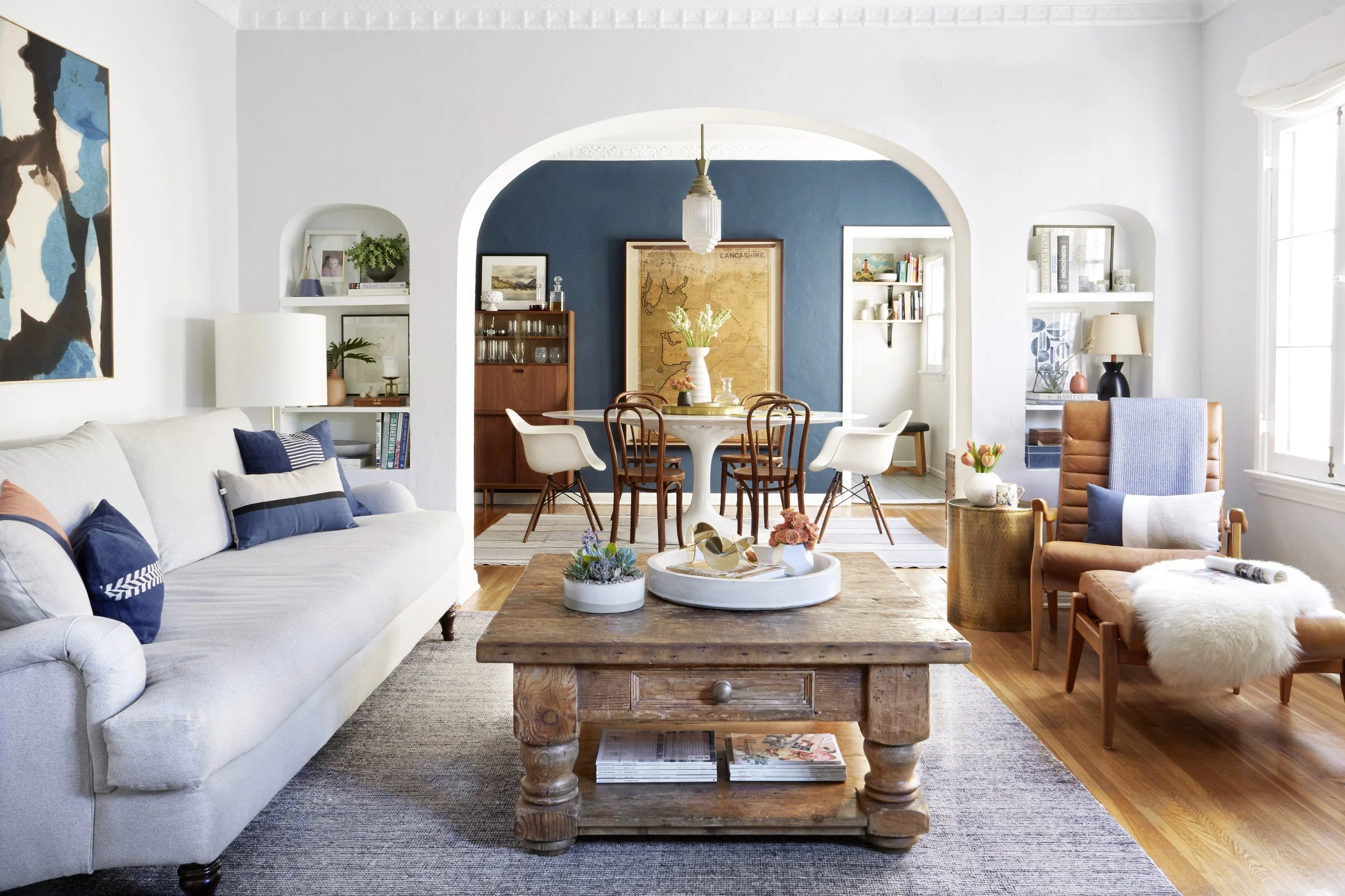 A living room with white walls and a gray sofa against the wall. A blue-painted dining room is in the background.