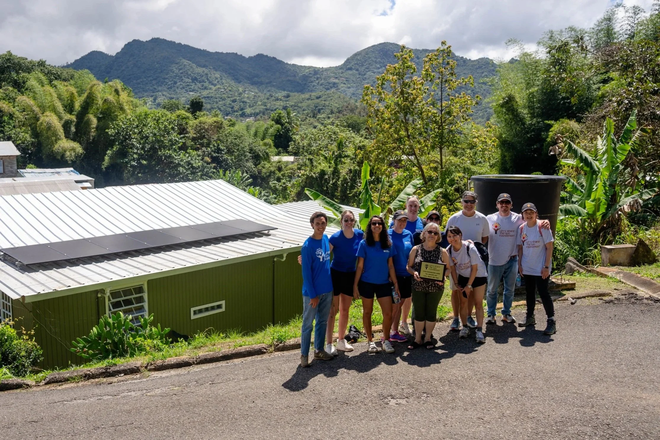 Wood Mackenzie & LSTS team alongside beneficiary after installation. Group stands in front of house with solar panels visible.