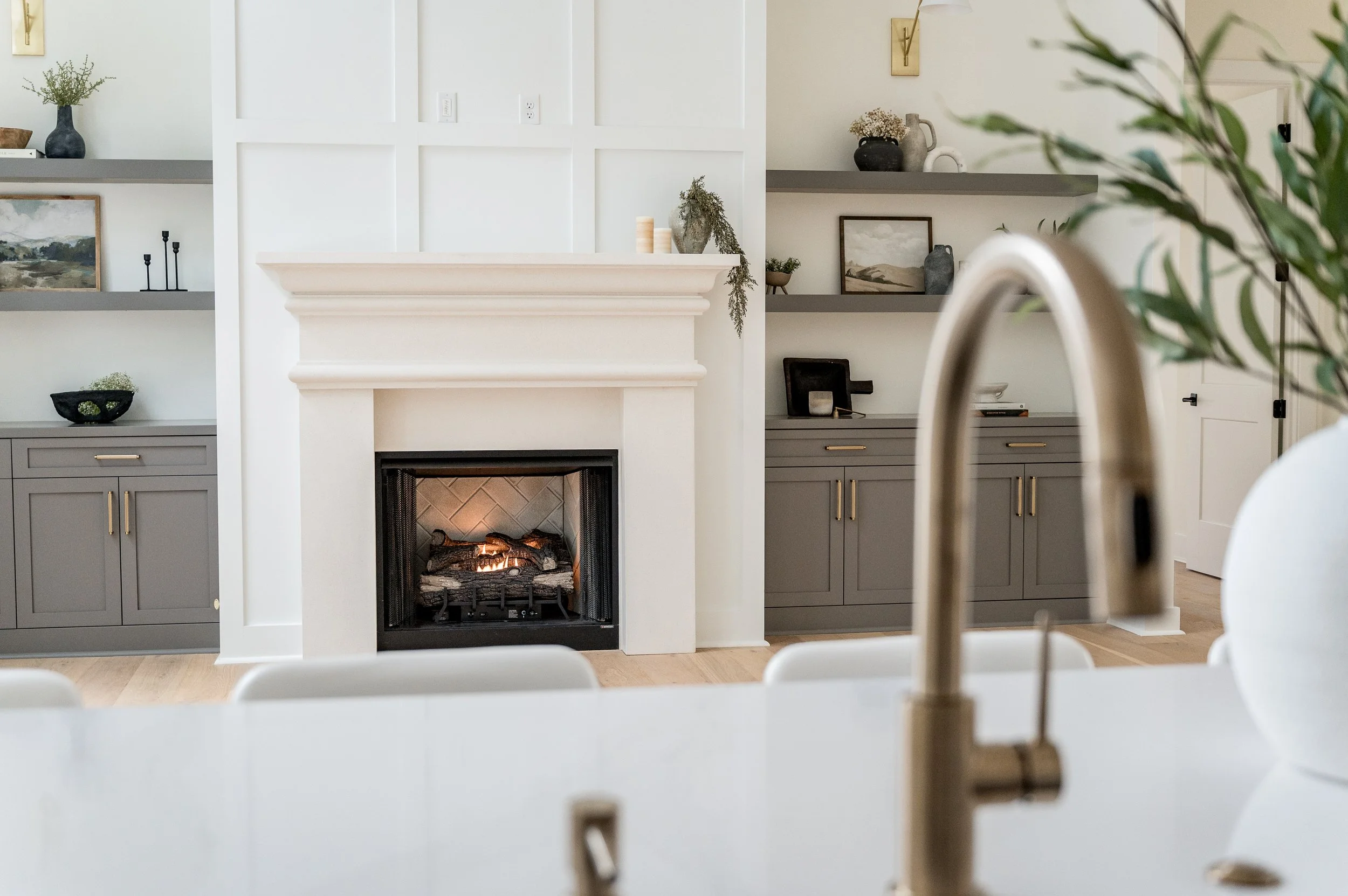 Modern living room with a fireplace, grey cabinets, and open shelves with decor items. A kitchen faucet is partially visible in the foreground.