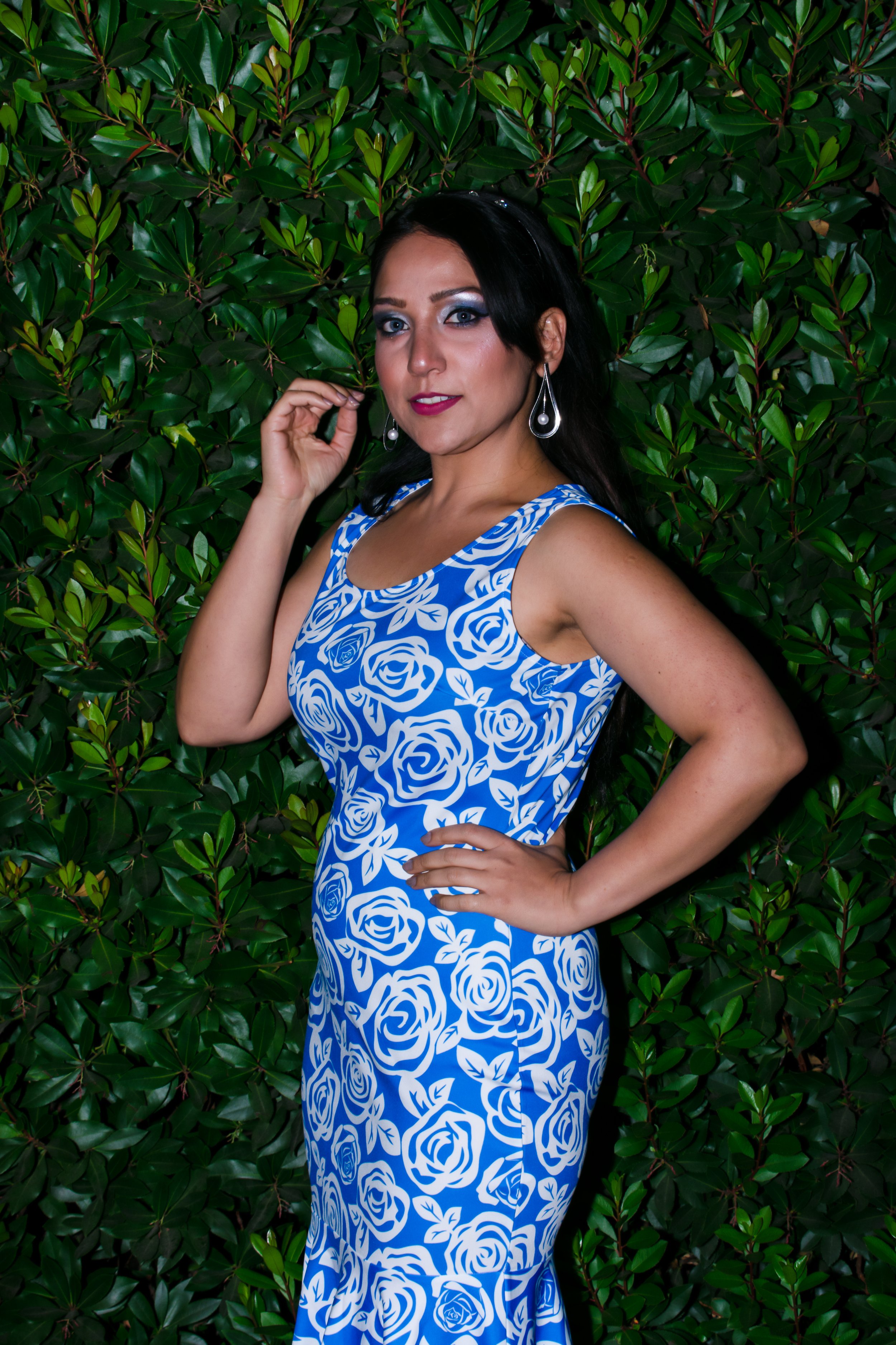 A woman with dark hair and makeup wearing a blue and white floral dress, silver earrings, standing against a green leafy background.