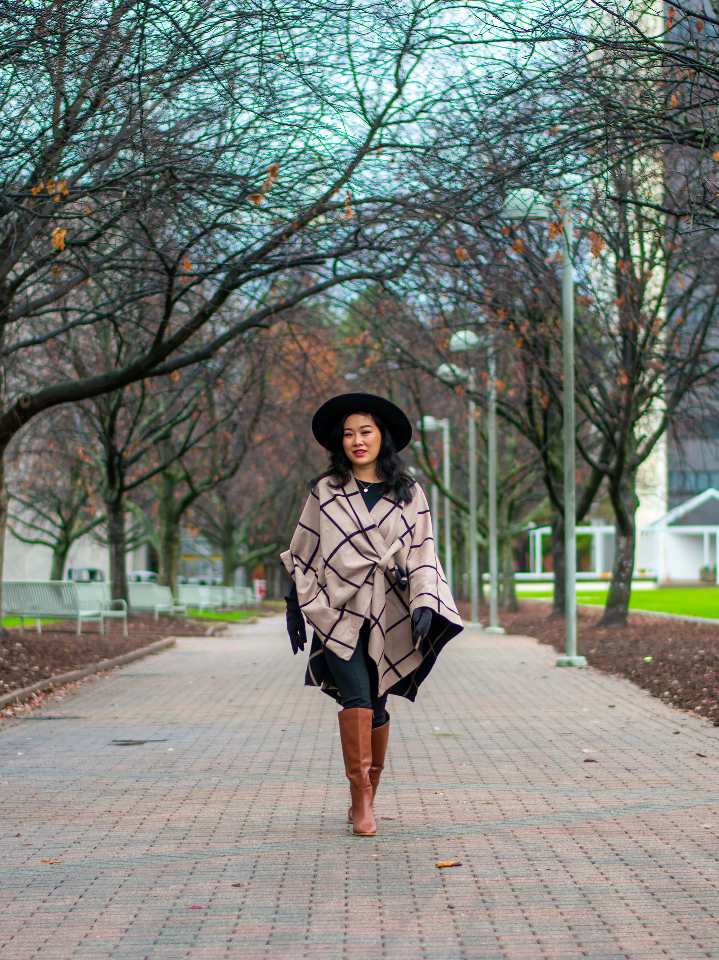 A woman walking on a paved pathway in a park lined with leafless trees, wearing a beige checkered coat, tall brown boots, black gloves, and a black hat, with a neutral expression.