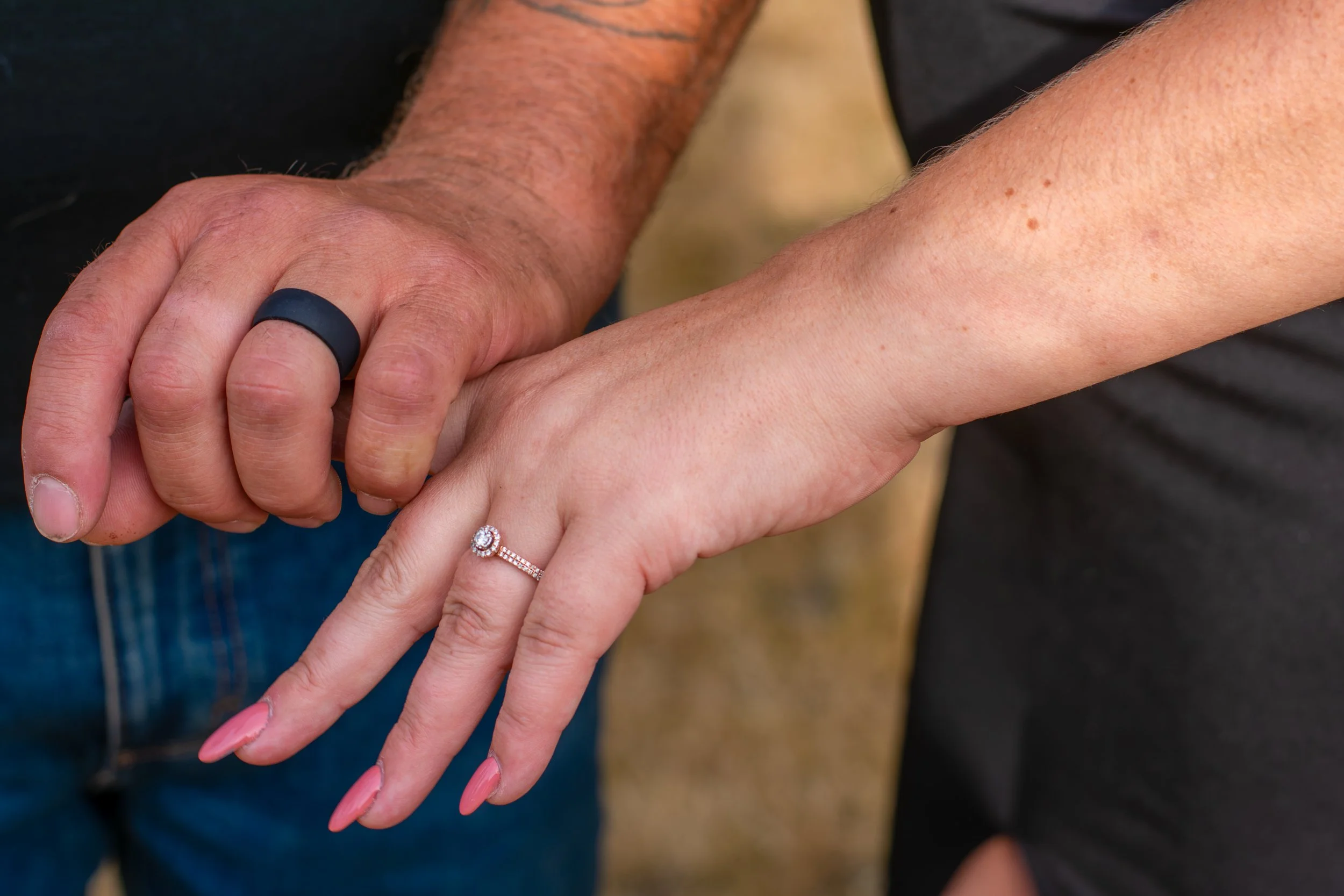 Close-up of a couple holding hands, showcasing a wedding ring on the woman's finger. The man's hand is larger with visible hair, and he wears a black ring. The woman's hand has a diamond engagement ring. Their hands are gently resting on each other, 