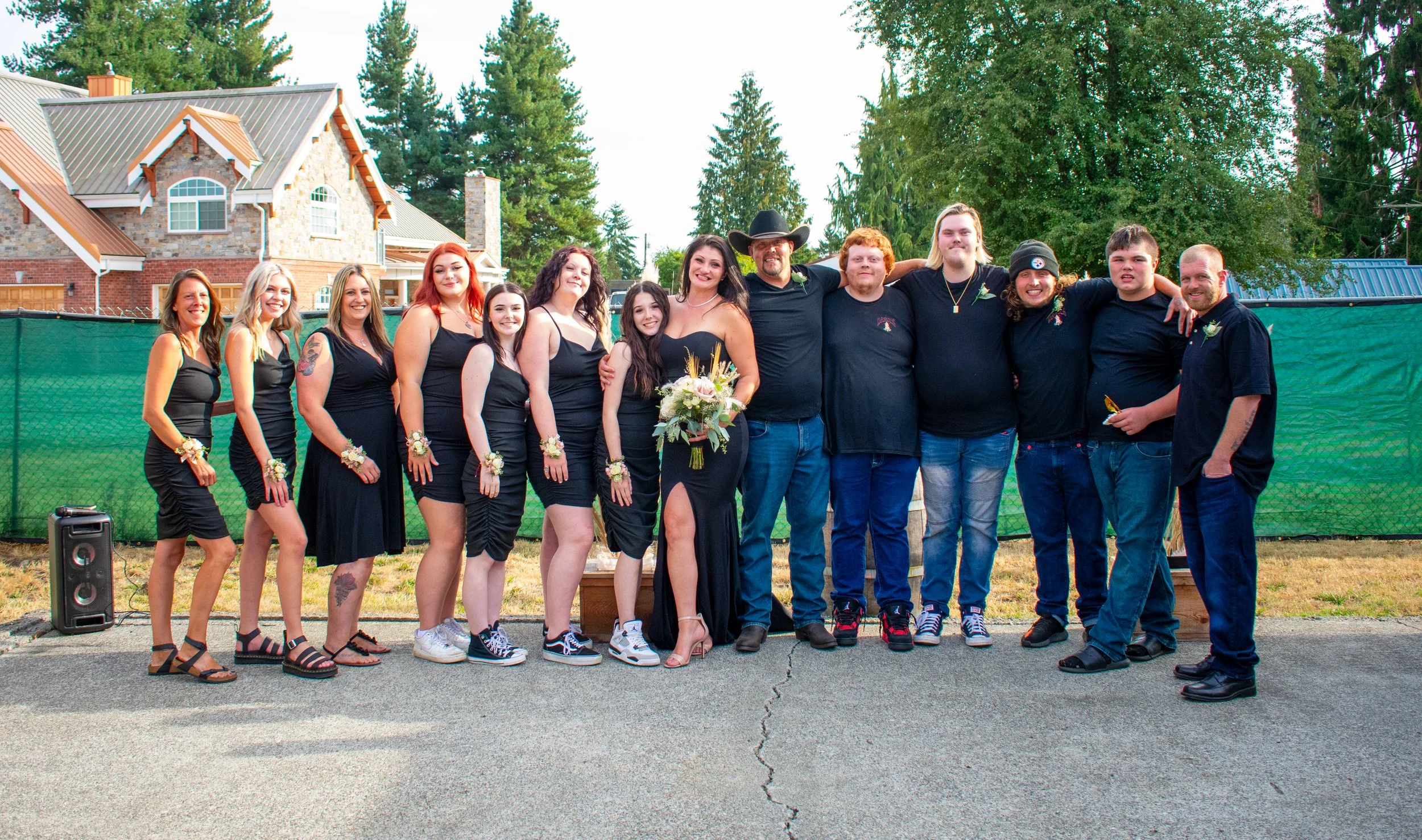 A group of sixteen people standing outdoors in front of a green fence and large house, smiling for a photo. The group includes women in black dresses and men in casual clothing, some wearing corsages or boutonnières, indicating a celebratory event su