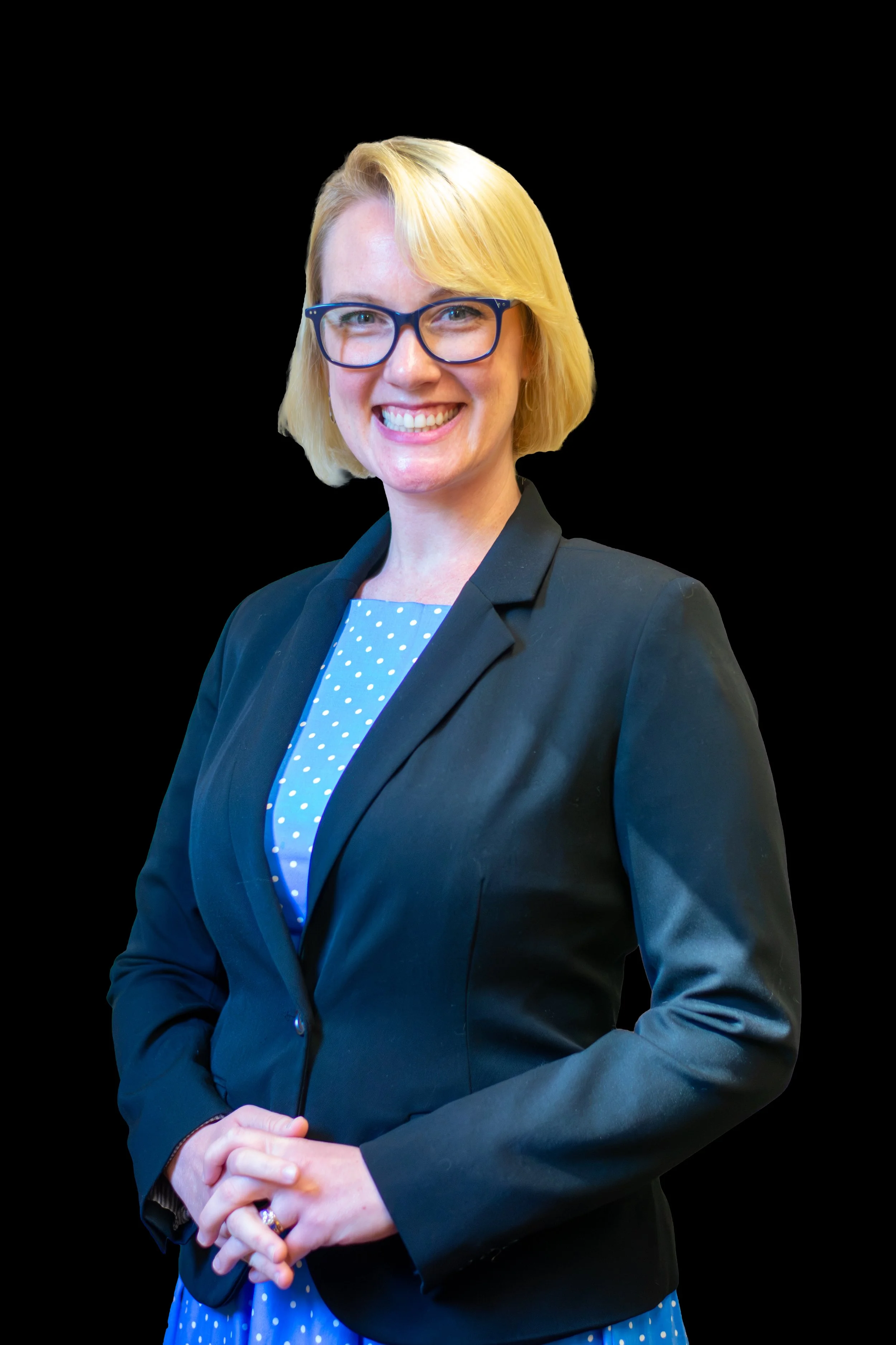 A woman with blonde hair, wearing glasses, a dark blazer, and a blue polka dot dress, smiling and standing with hands clasped in front against a black background.