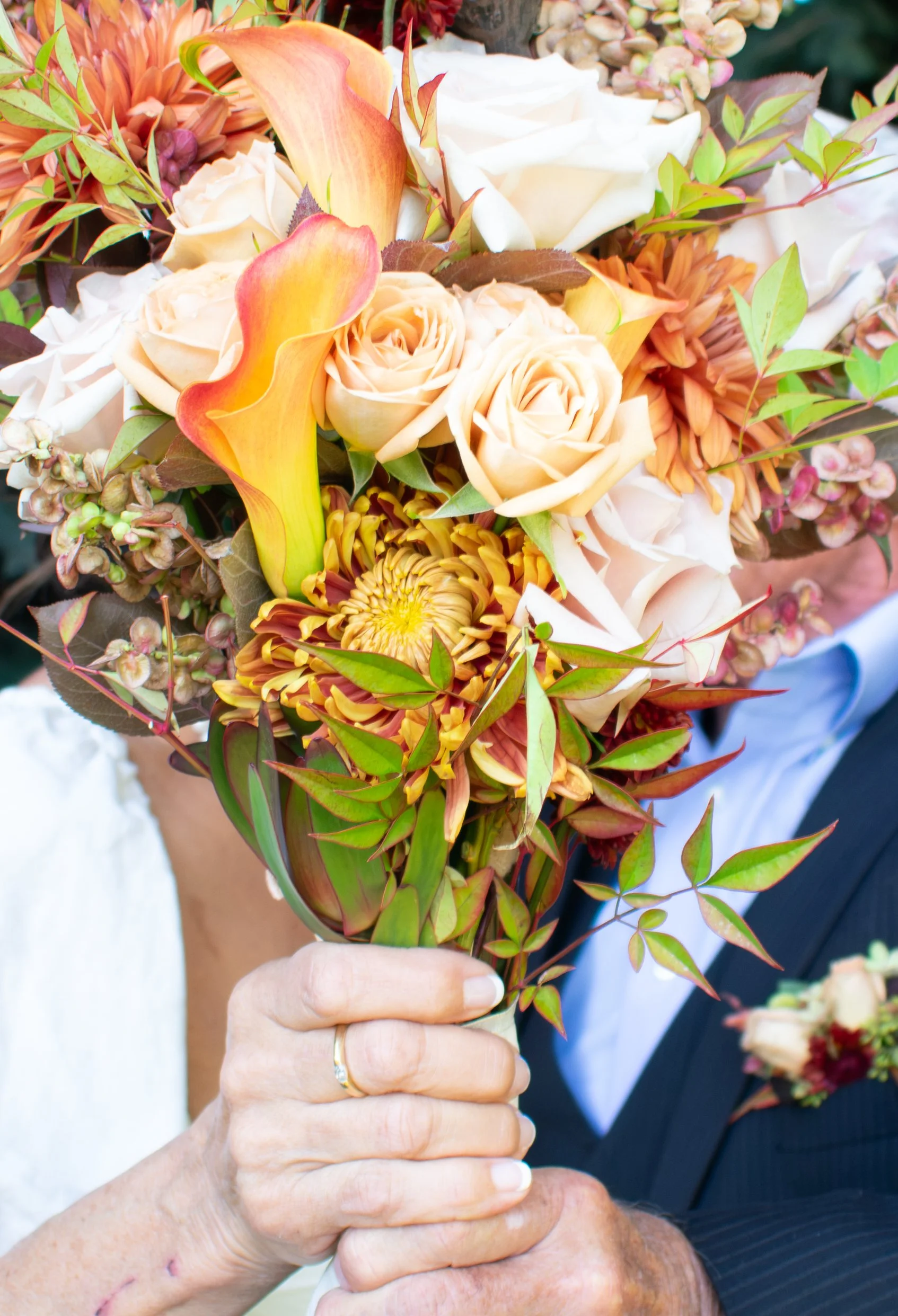 Close-up of hands holding a bouquet of flowers, including roses, calla lilies, and other colorful blooms, with a person in a dark suit in the background.