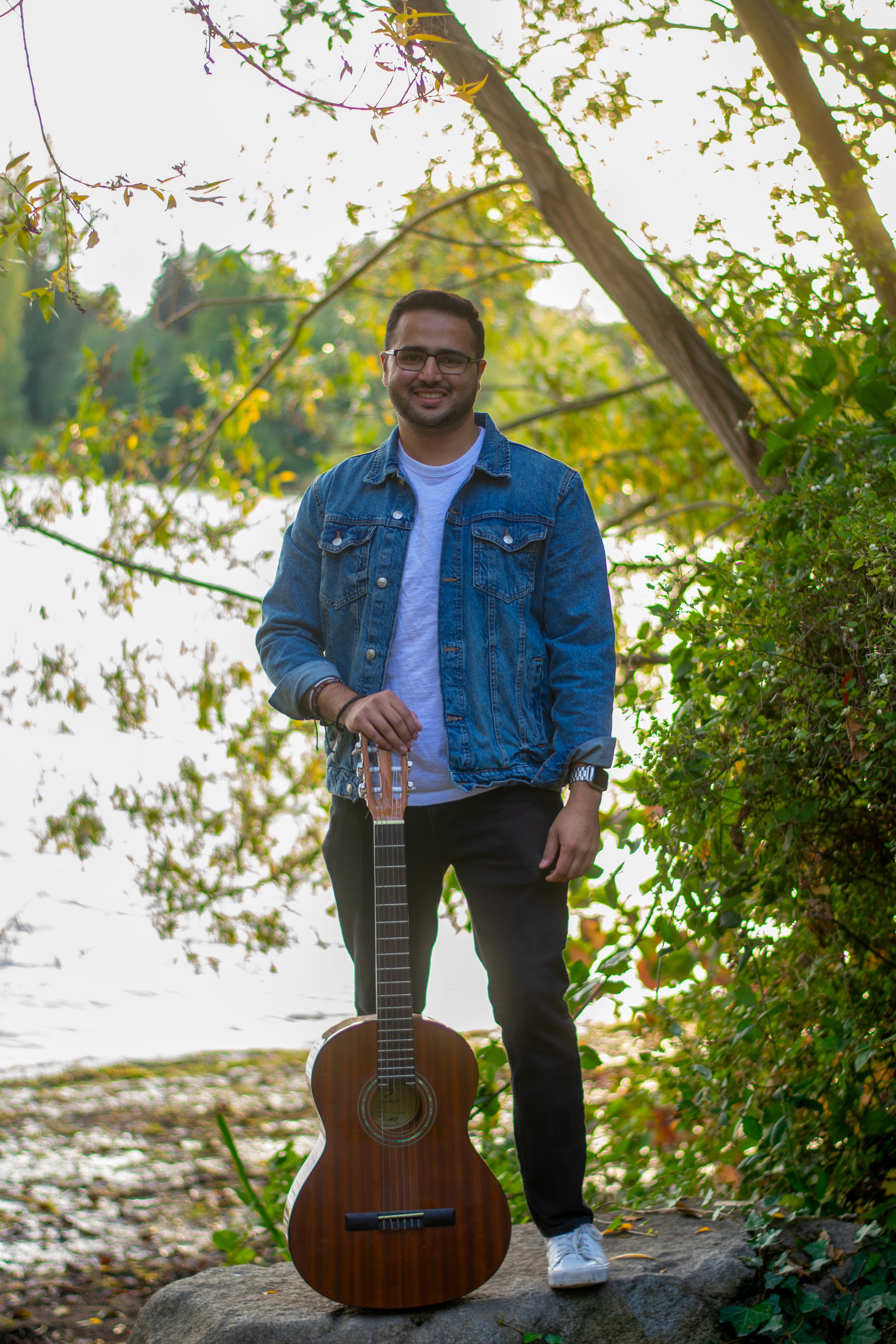 A man standing on a rock outdoors holding a guitar, surrounded by trees and foliage with sunlight filtering through.