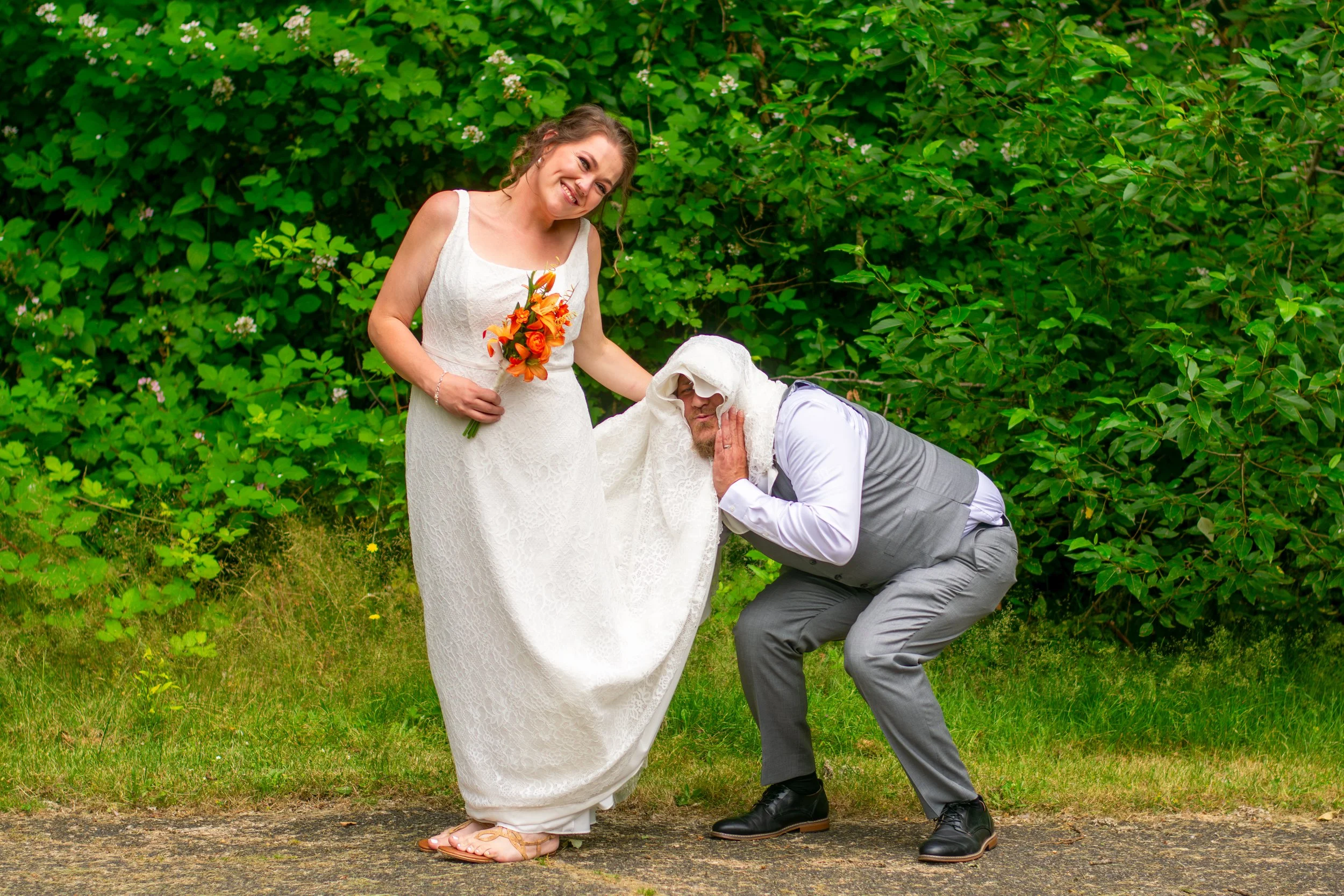 A woman in a white dress holding a bouquet of orange flowers is standing outdoors on a green grassy area with bushes in the background. A man in a gray vest and pants, with a white shirt, is kneeling and kissing her wedding dress on the leg while wea