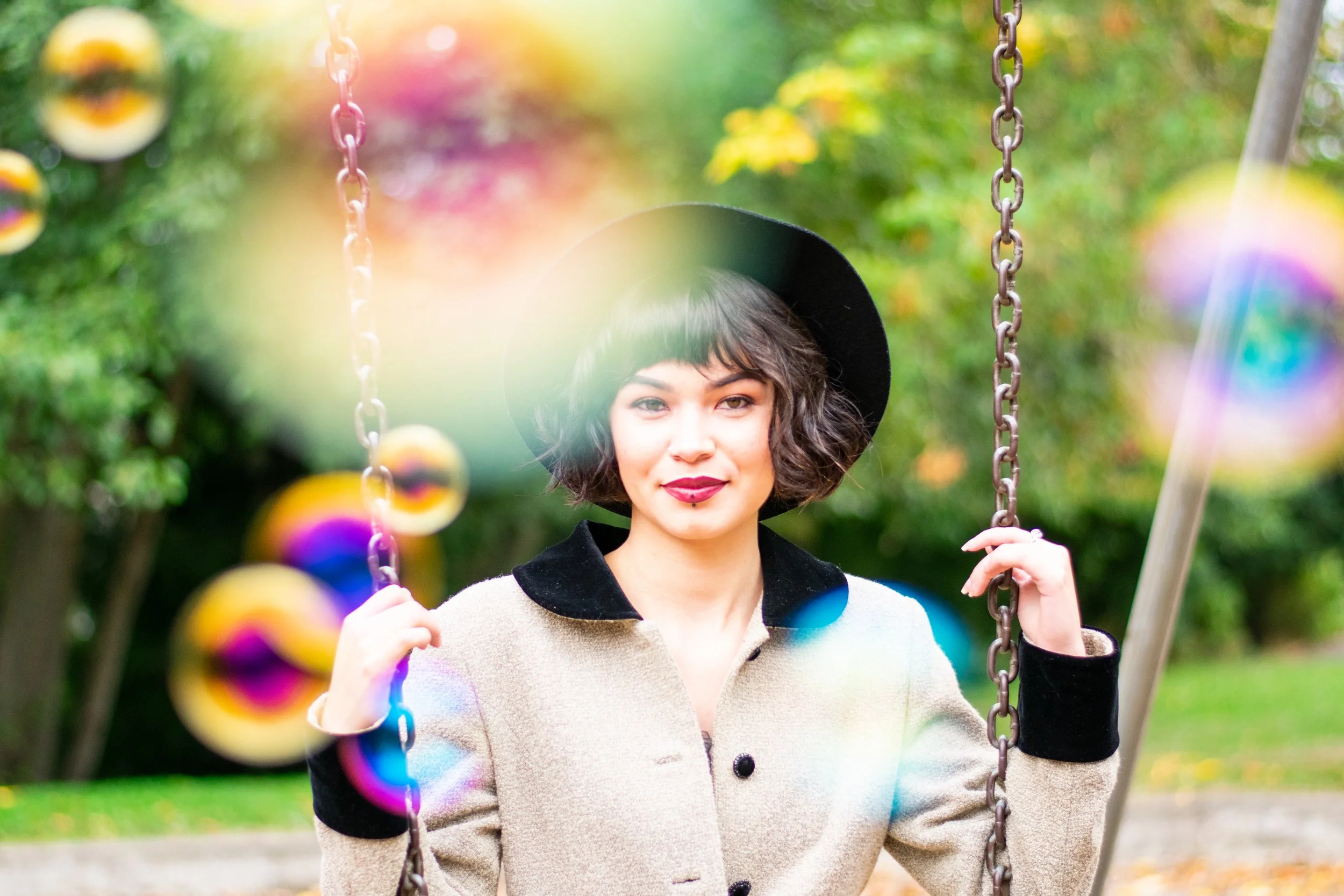 A woman with dark curly hair, wearing a beige coat with black cuffs and a black hat, is sitting on a swing in a park. There are colorful spinning tops moving in front of her, and trees with green leaves in the background.