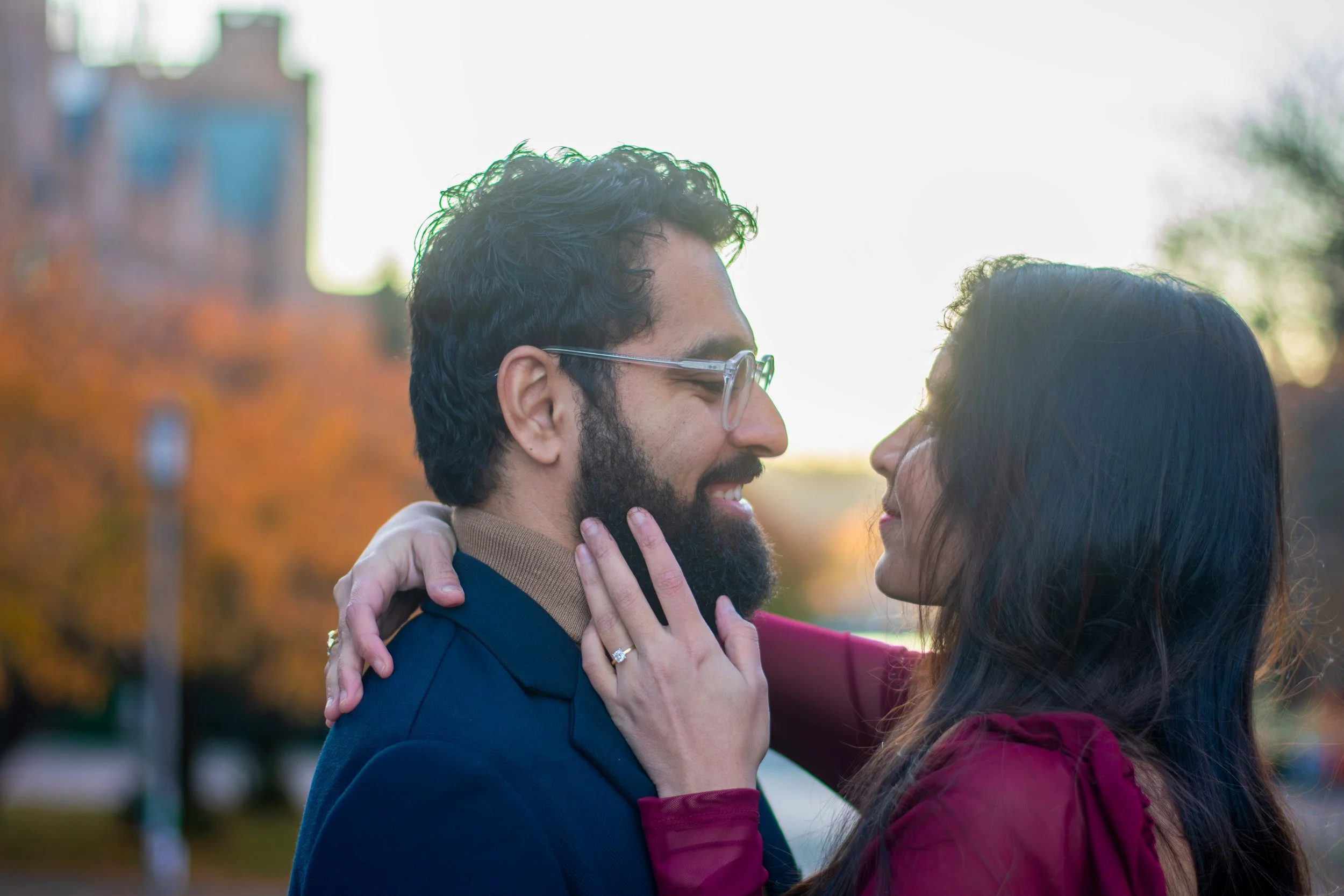 A man and woman closely facing each other outdoors in fall. The woman’s hand is on the man’s face, and they are smiling. The background shows trees with orange leaves and a blurred building.