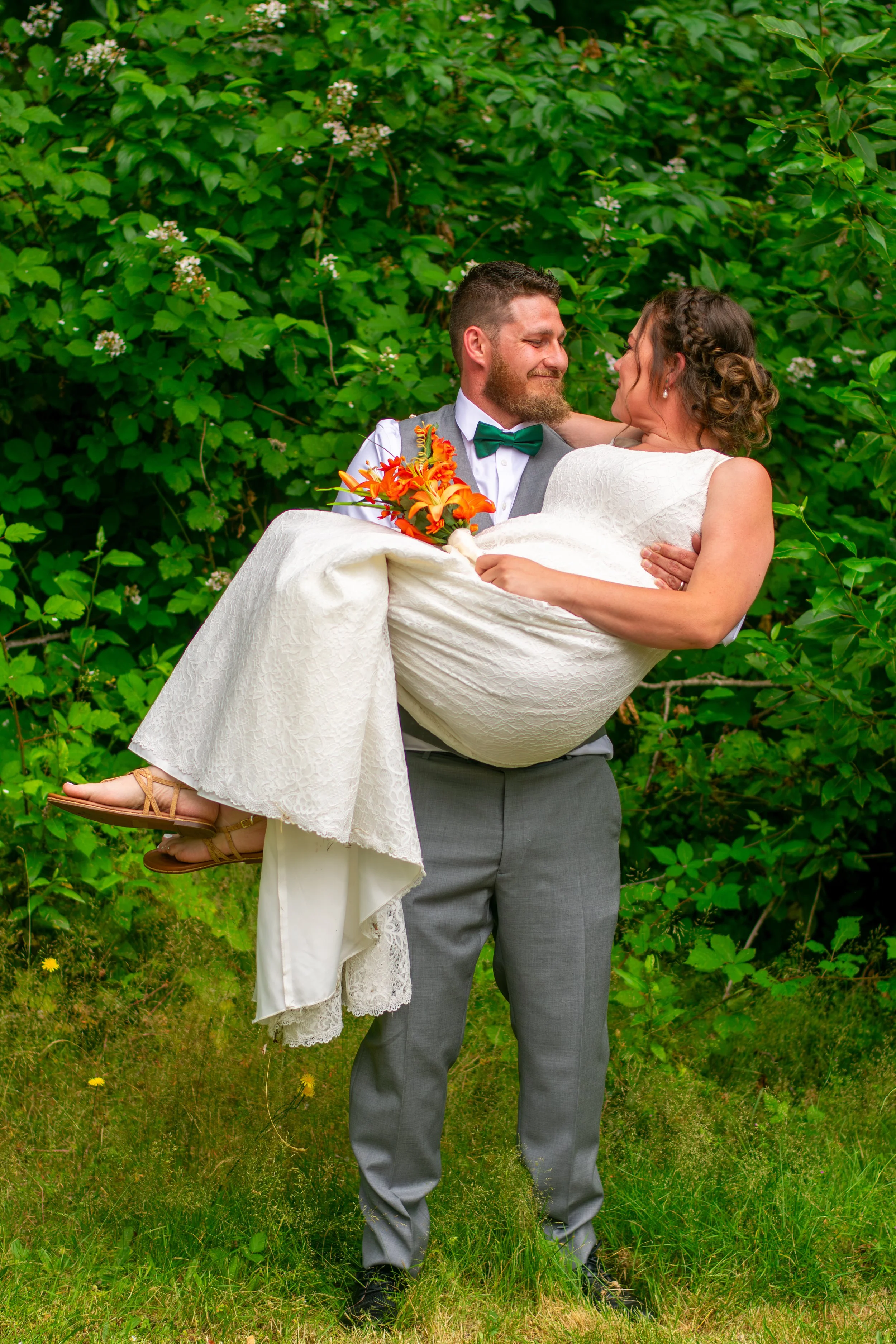 A groom in a gray suit with a green bow tie holding a bride in a white dress and heels, with a floral bouquet, standing outdoors in front of green foliage.