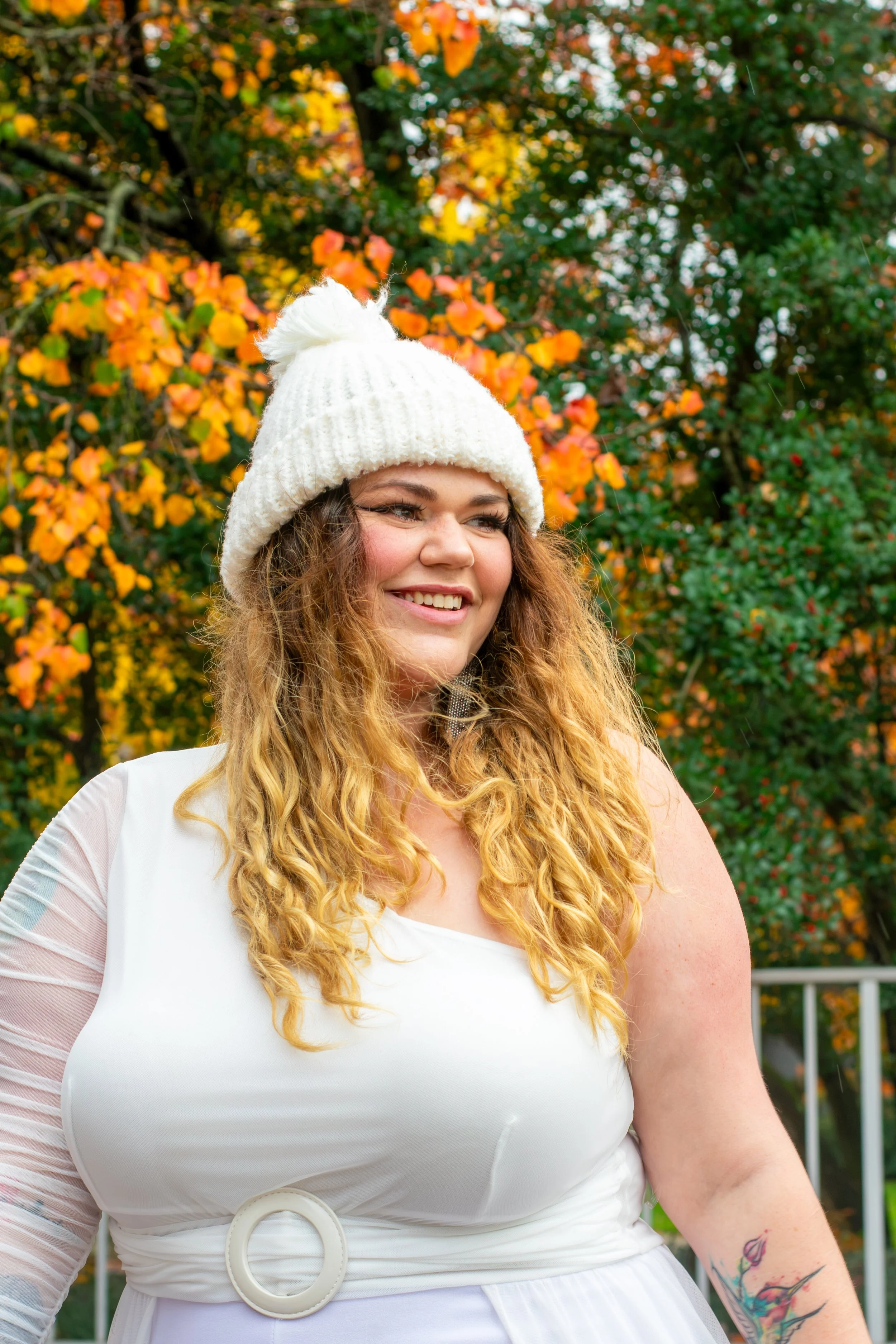 A woman with long, curly red hair wearing a white knit beanie with a pom-pom. She is outdoors, smiling, with colorful autumn leaves in the background. She wears a white top with mesh sleeves and has a colorful tattoo on her arm.