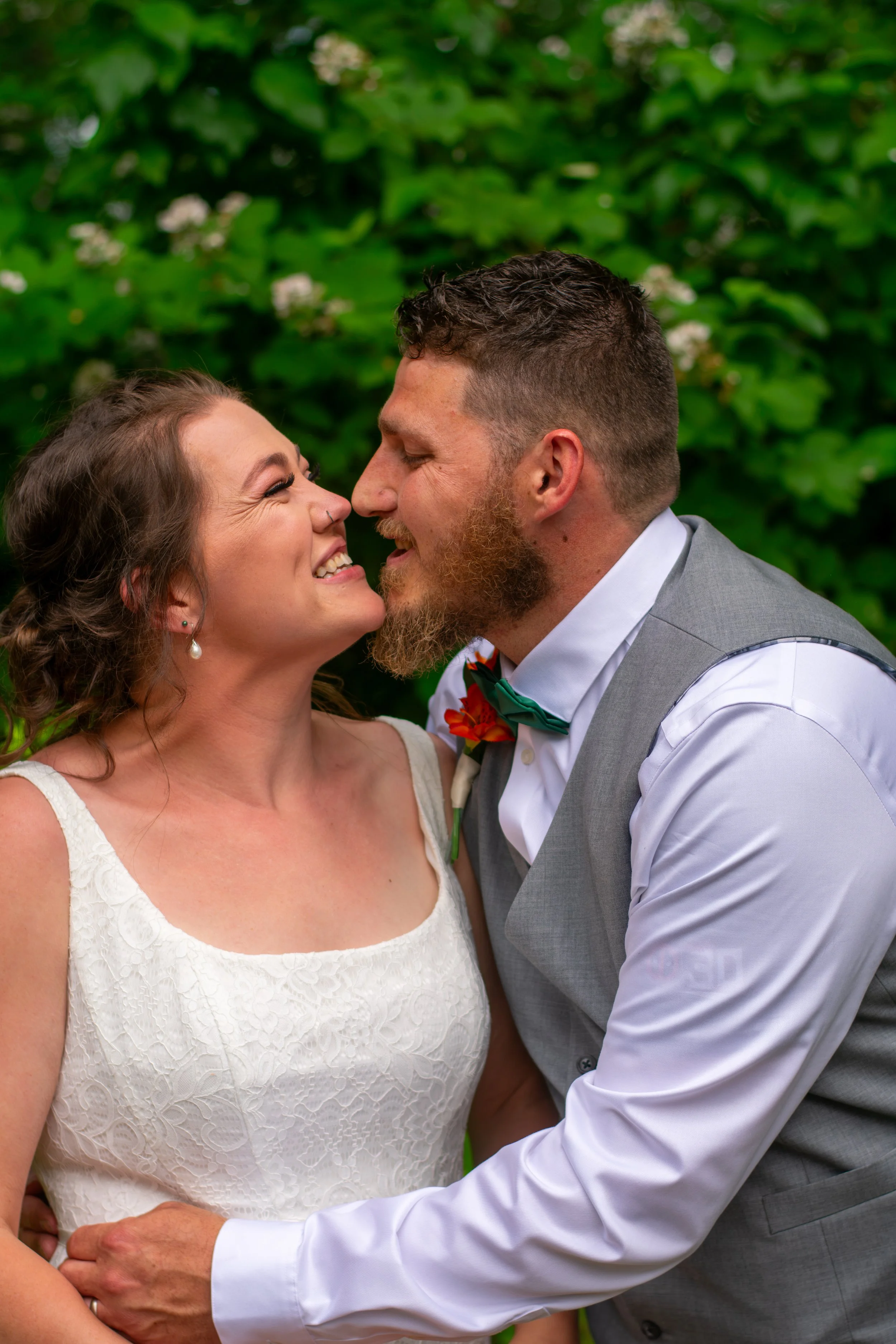 A couple on their wedding day, smiling and about to kiss, outdoors with green foliage in the background. The bride is wearing a white lace dress and pearl earrings, while the groom is in a light gray vest, white shirt, and a green bow tie with a flor