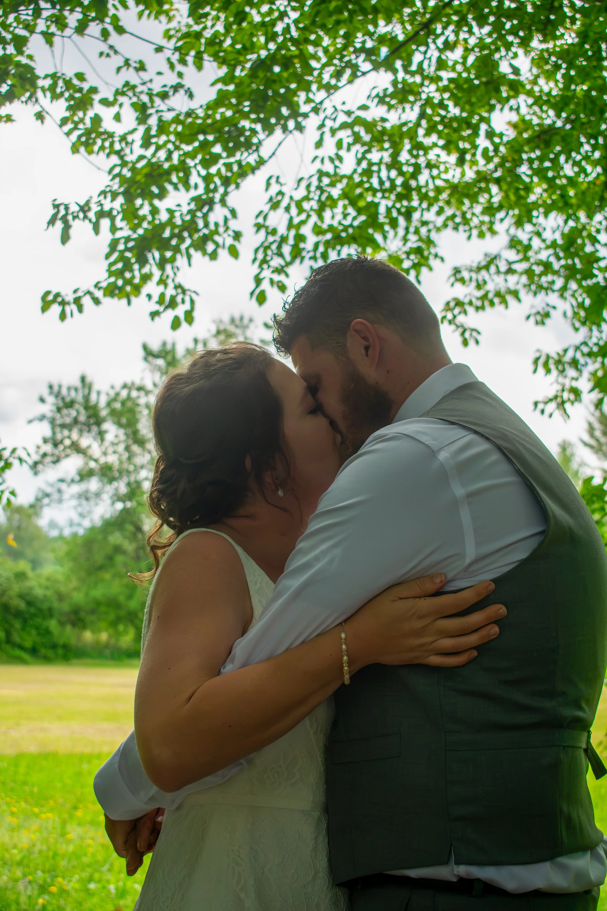A couple dressed in wedding attire sharing a kiss outdoors under a large tree with green leaves, with a grassy field and yellow flowers in the background.