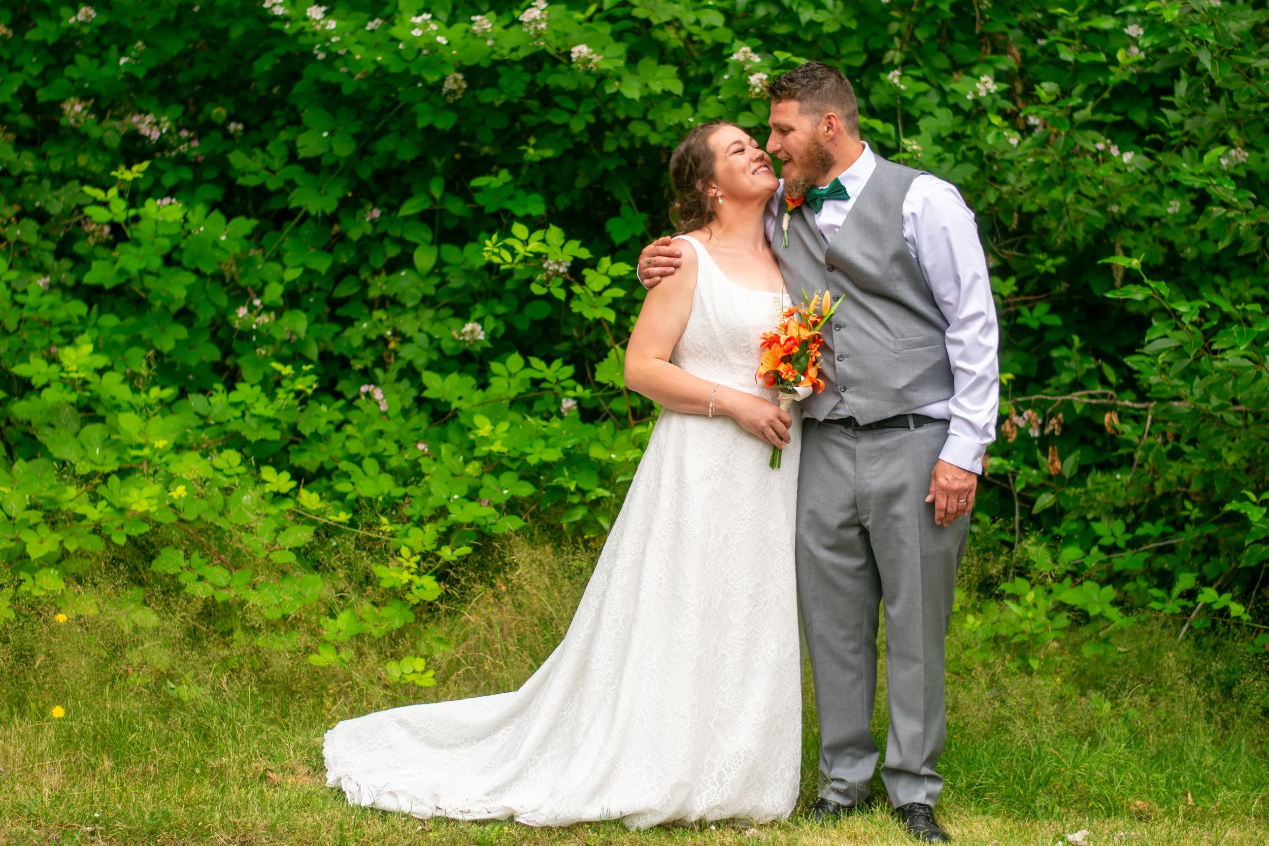 A bride and groom on their wedding day, standing close in front of green foliage with white flowers. The bride wears a white lace wedding dress and holds a bouquet of orange and yellow flowers. The groom wears a gray suit with a white shirt, a green 