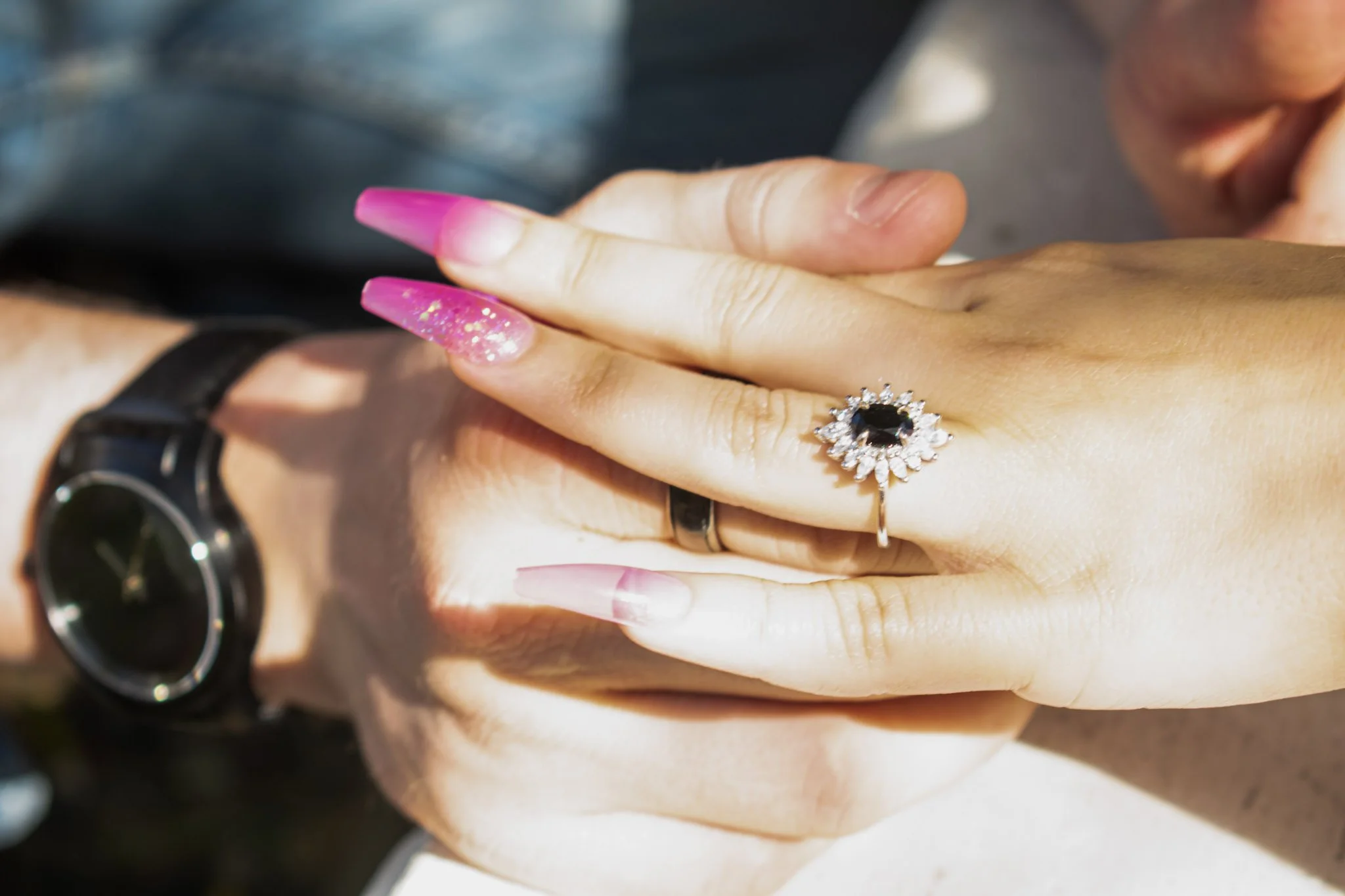 Close-up of a person's hand with pink and glittery nails, wearing a silver ring with a black gemstone, and a black wristwatch. The background is blurred.
