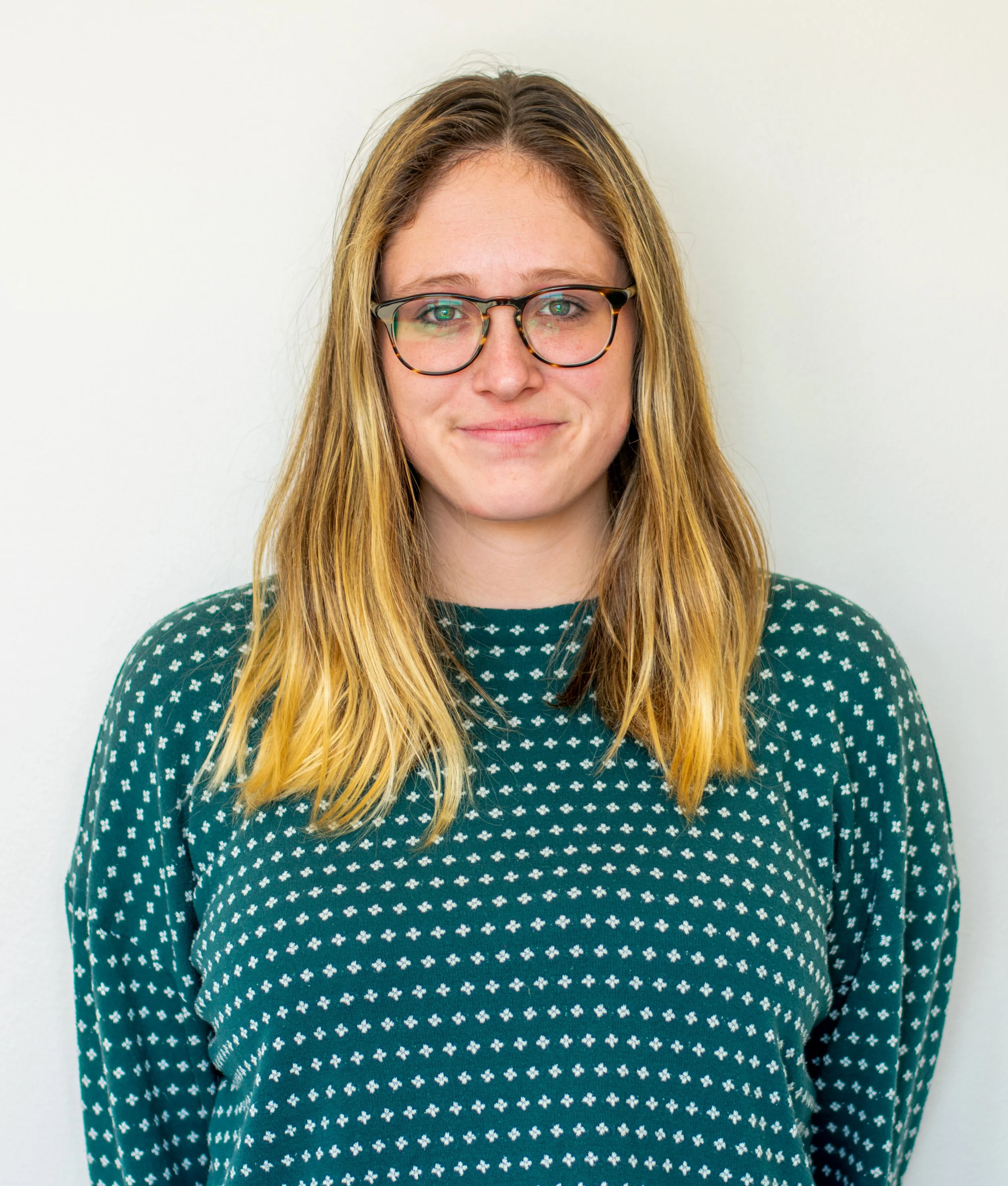 A woman with long blonde hair, green eyes, and glasses, wearing a green sweater with white cross patterns, standing against a plain white background.