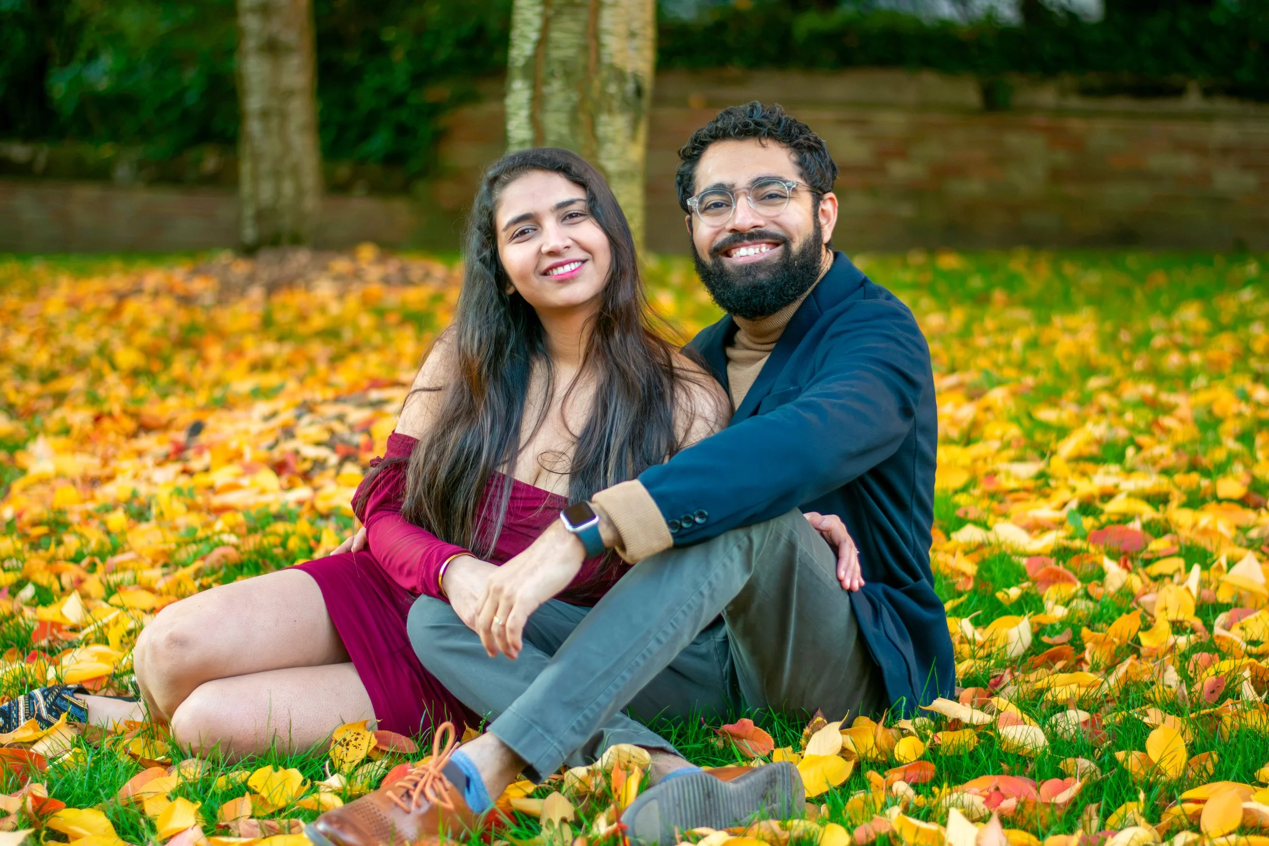 A smiling young couple sitting on a bed of colorful autumn leaves in a park, with trees and a brick wall in the background.