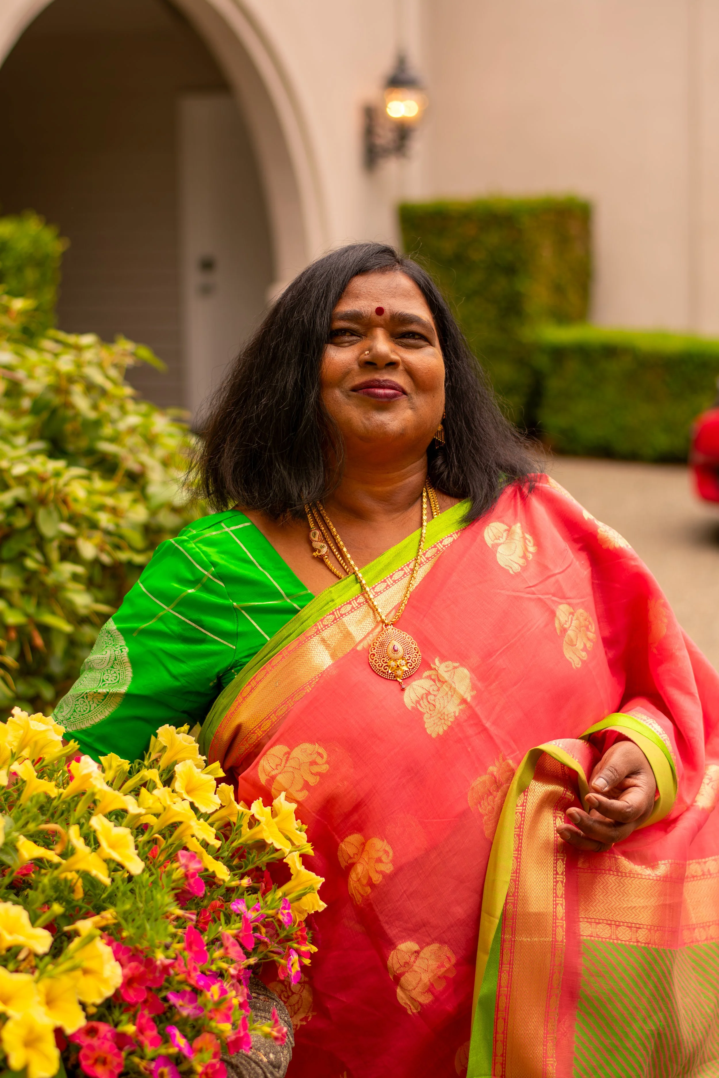 A woman dressed in a pink and green traditional Indian saree, standing outdoors near colorful flowers.