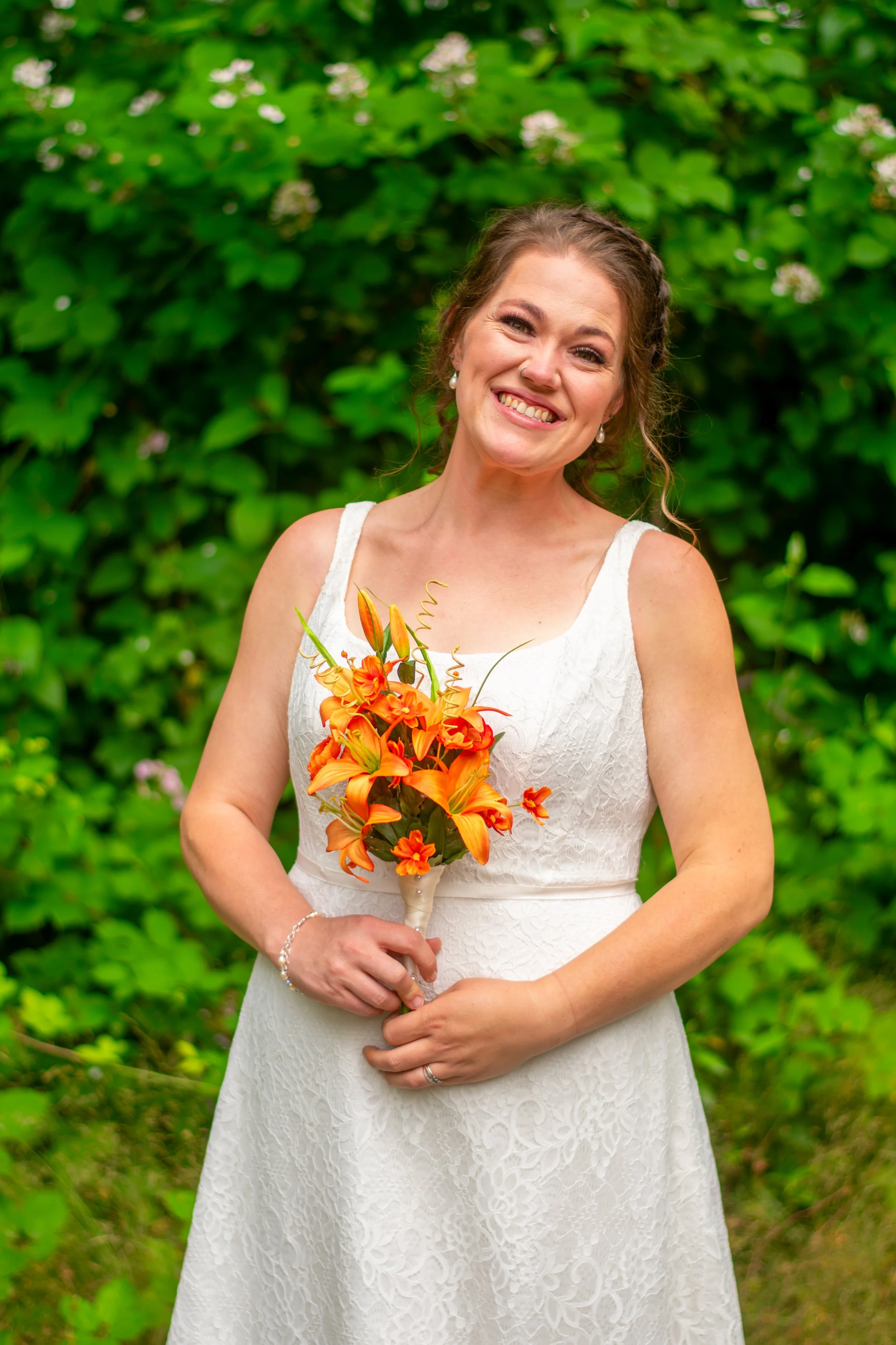 A smiling woman in a white wedding dress stands outdoors, holding a bouquet of orange lilies and other flowers, with a background of lush green foliage.