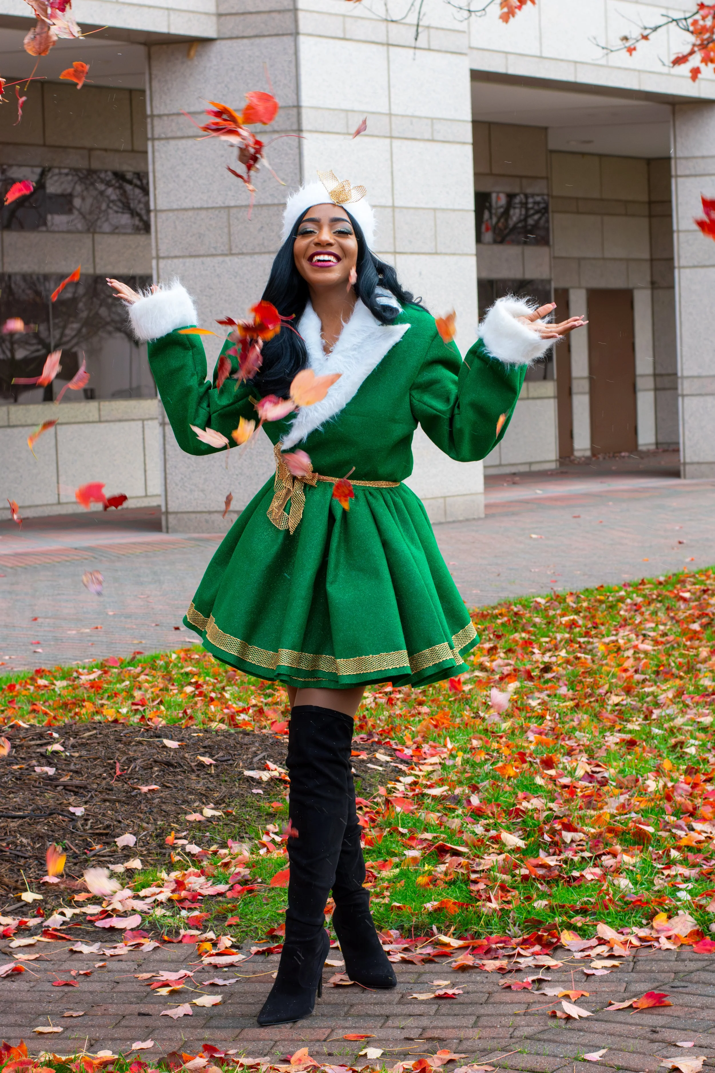 A woman wearing a green holiday dress and black boots standing outdoors on a brick sidewalk, surrounded by scattered autumn leaves, smiling and throwing leaves in the air.