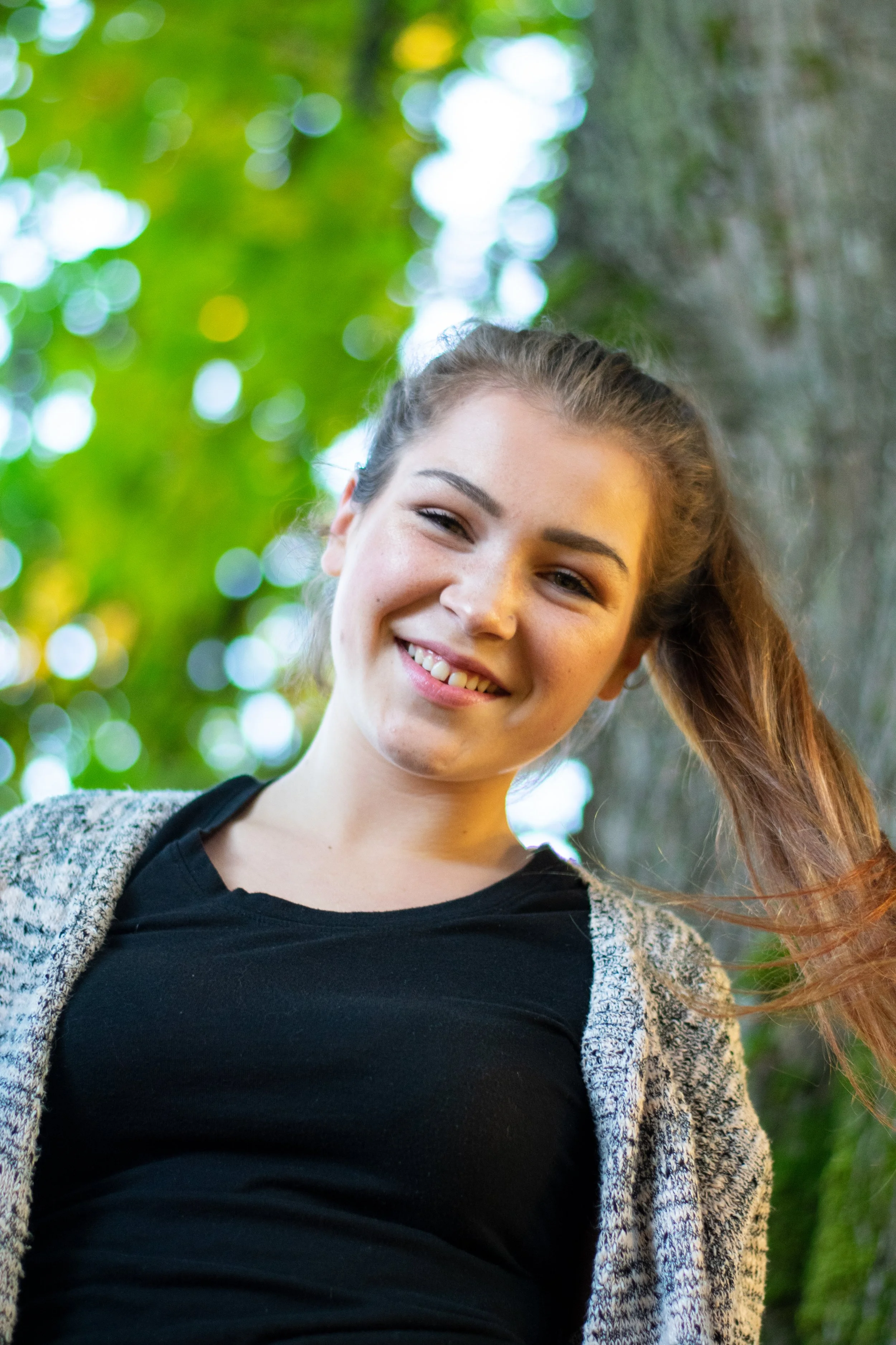 A young woman with long brown hair smiling outdoors, with green blurred trees in the background.
