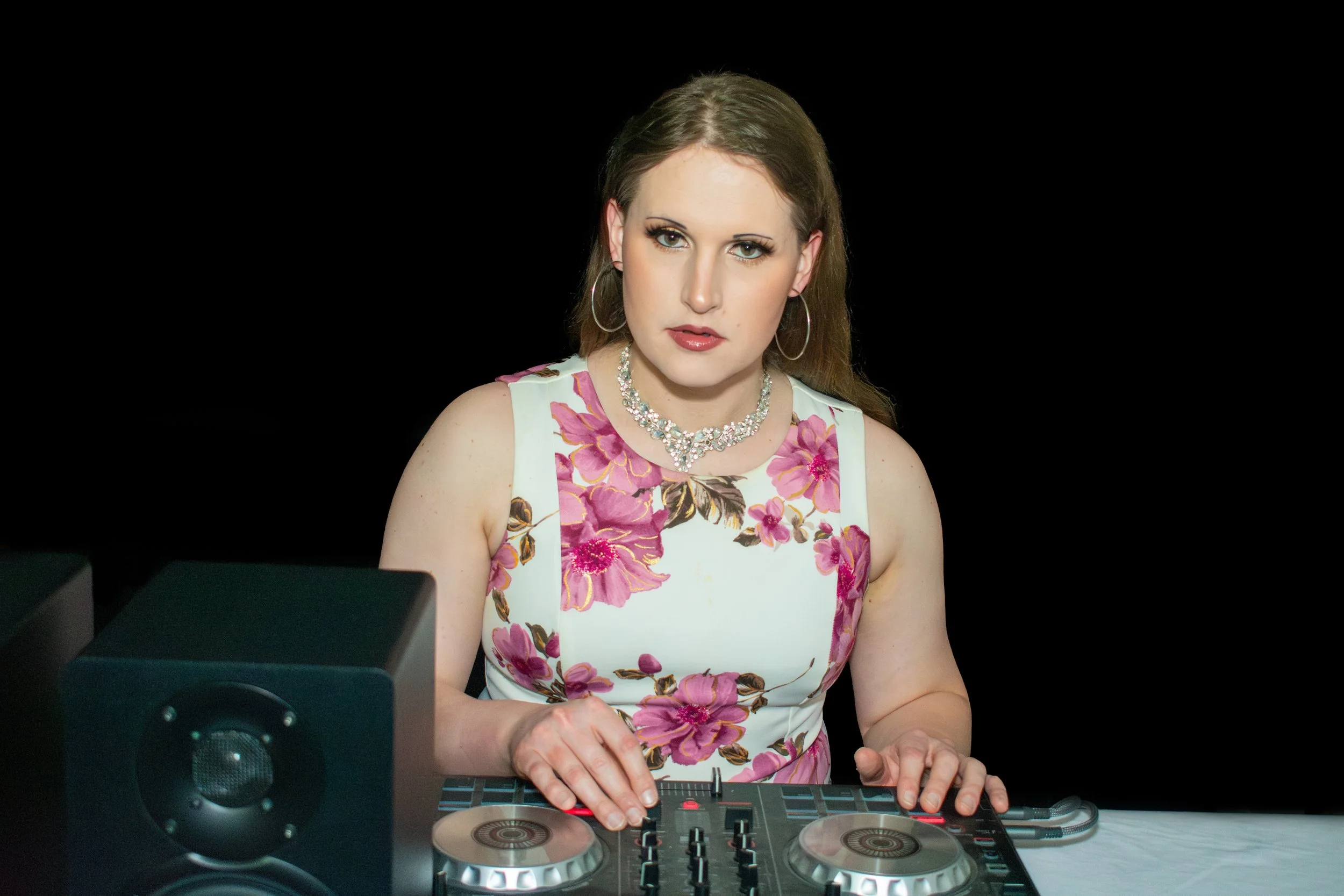 A woman in a floral dress is DJing with a controller, wearing large hoop earrings and a necklace.