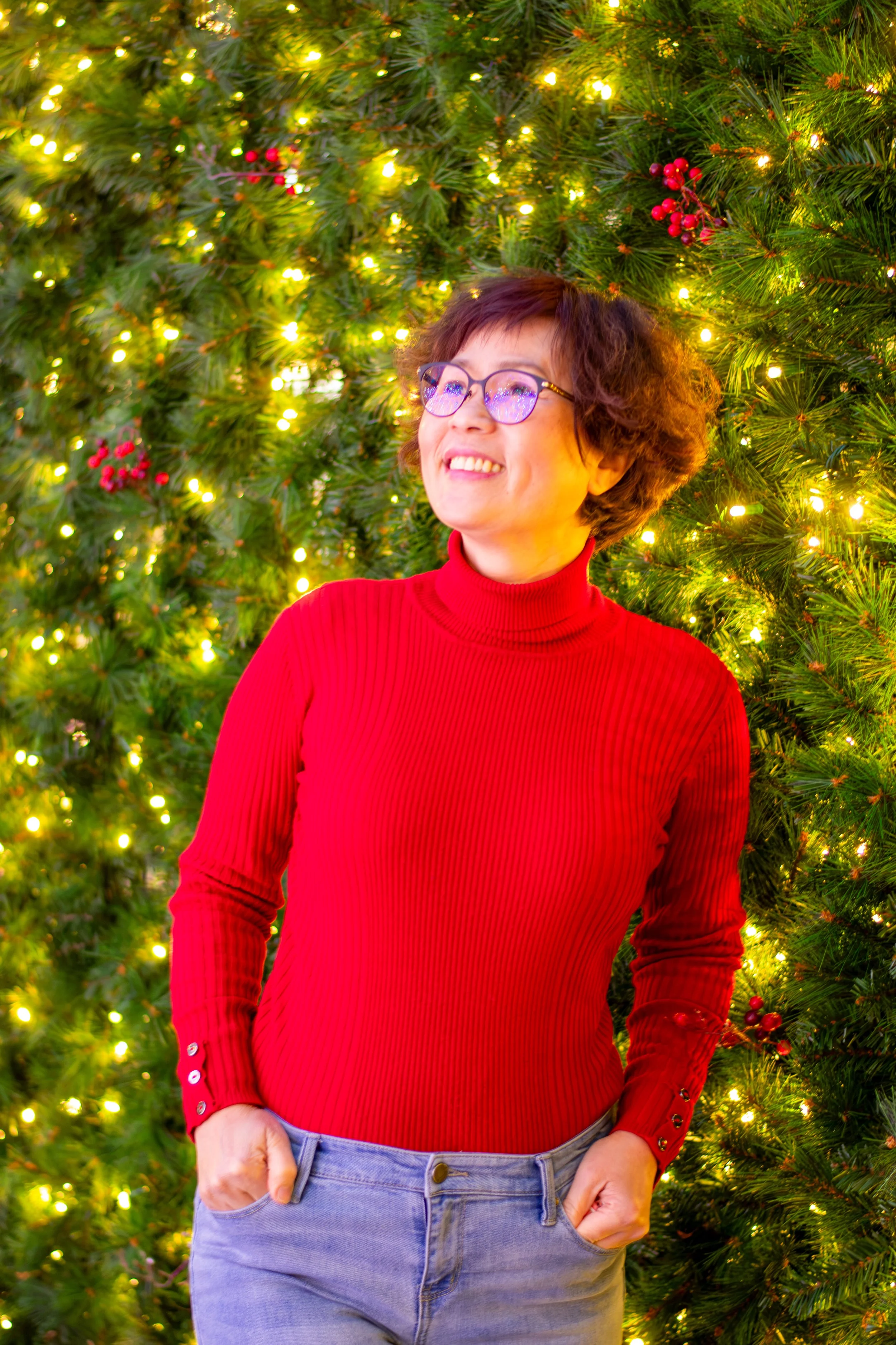 A woman smiling and wearing glasses, dressed in a red turtleneck sweater and light-colored jeans, standing in front of a decorated Christmas tree with green branches and yellow lights.
