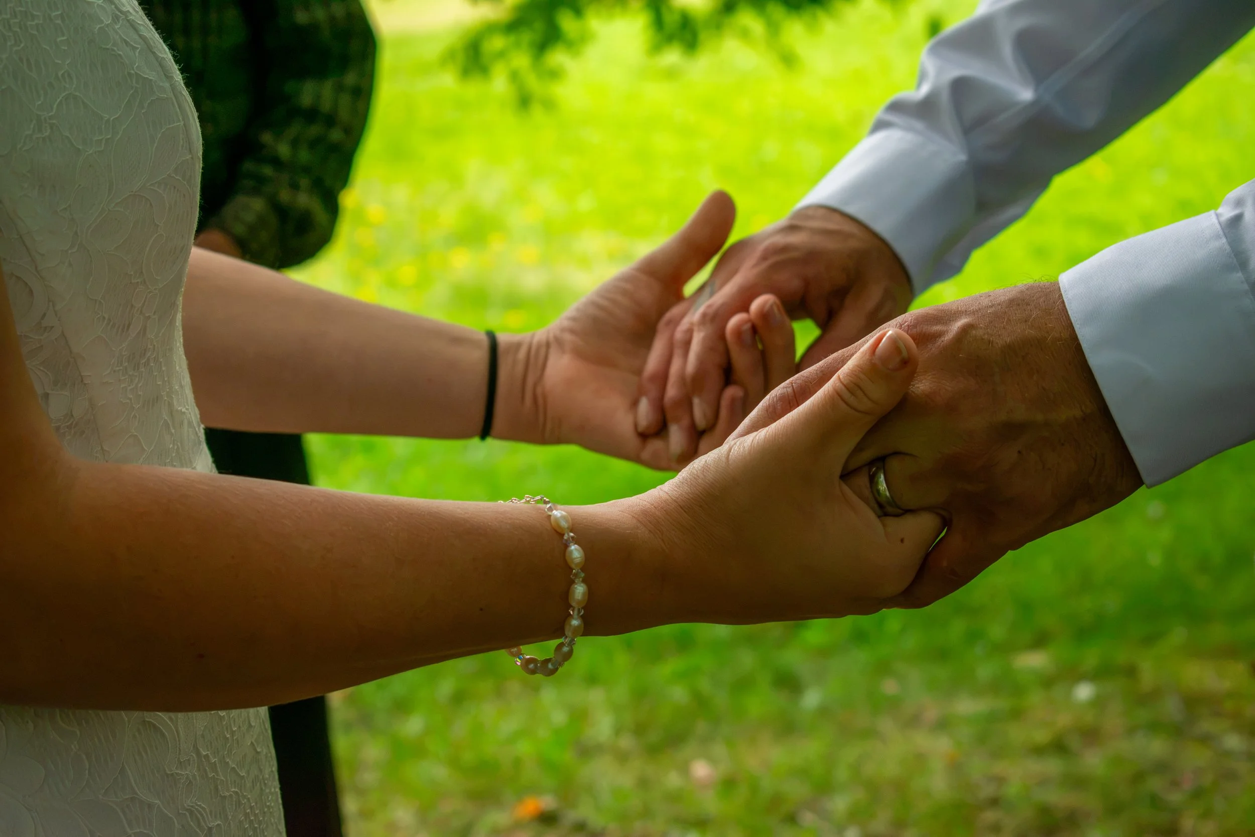 A couple holding hands outdoors with a green background, the woman wearing a pearl bracelet and the man wearing a wedding ring.