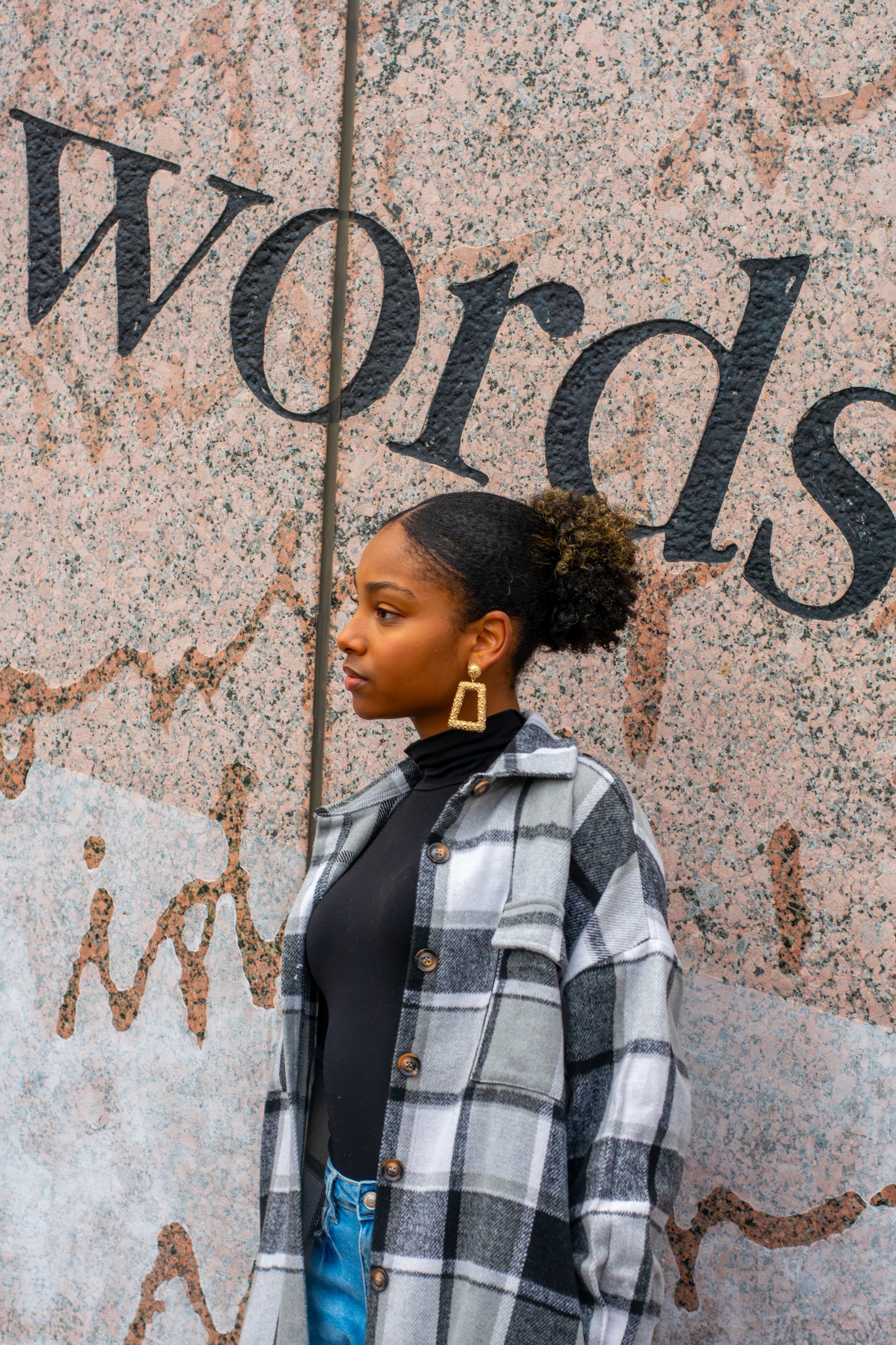 A young woman with curly hair in a bun, wearing large gold earrings, a black top, and a gray plaid coat, standing against a granite wall with the word "Wood" partially visible.