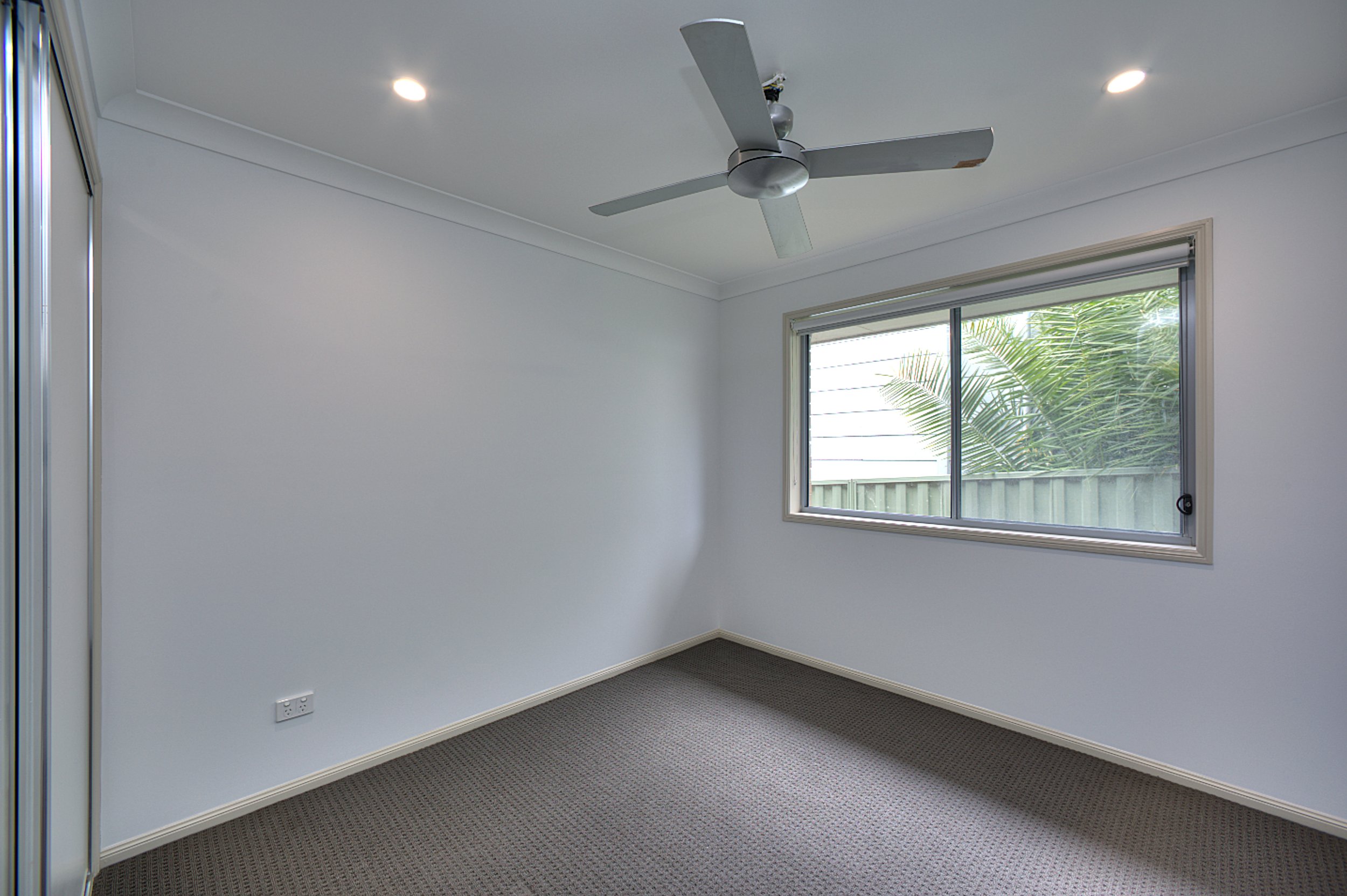 Empty bedroom with white walls, brown carpet, window with plants outside, ceiling fan, and recessed lighting.