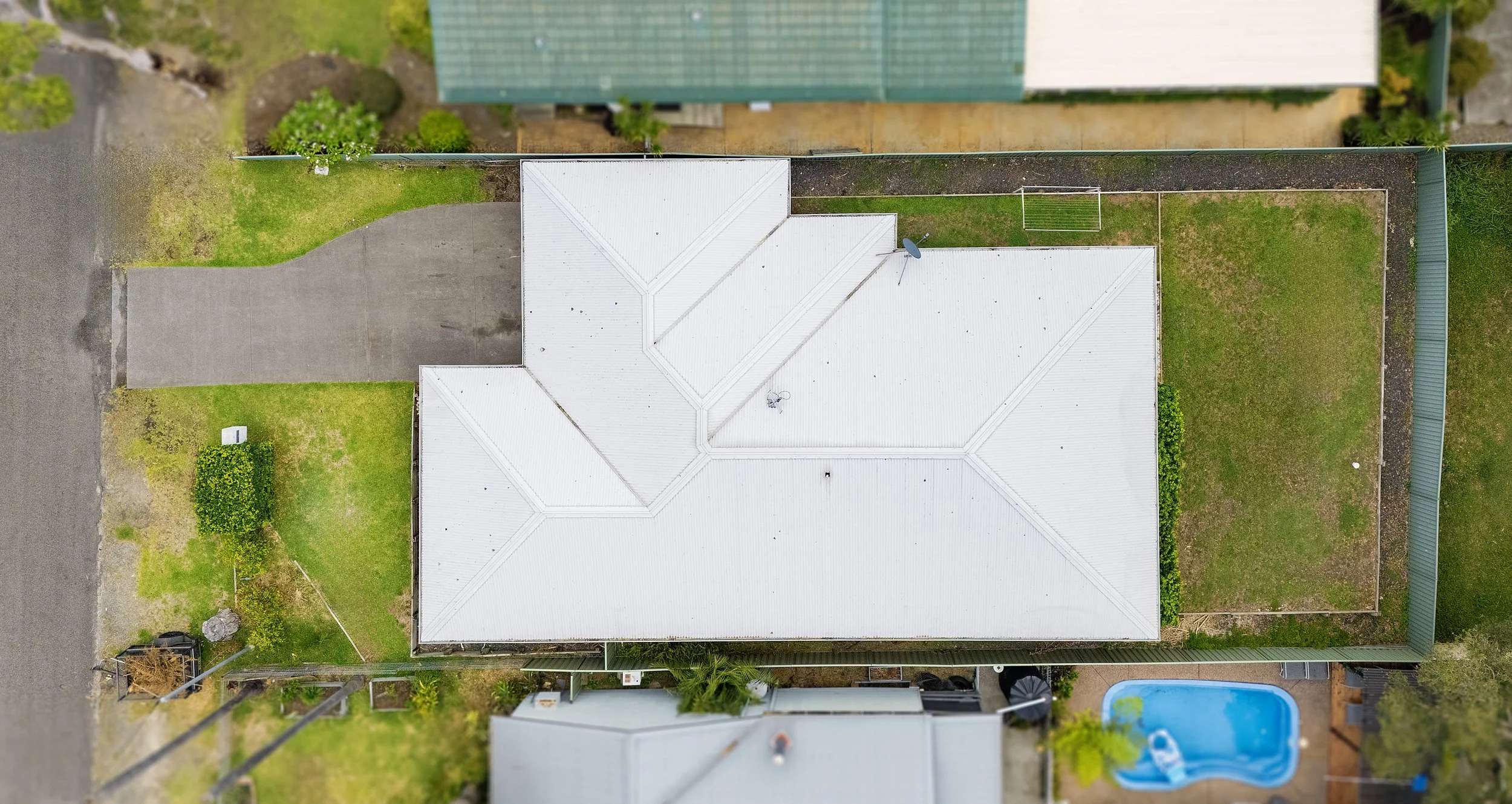 Aerial view of a house with a white roof, surrounded by green lawn, a driveway, a backyard with a small swimming pool, and neighboring houses.