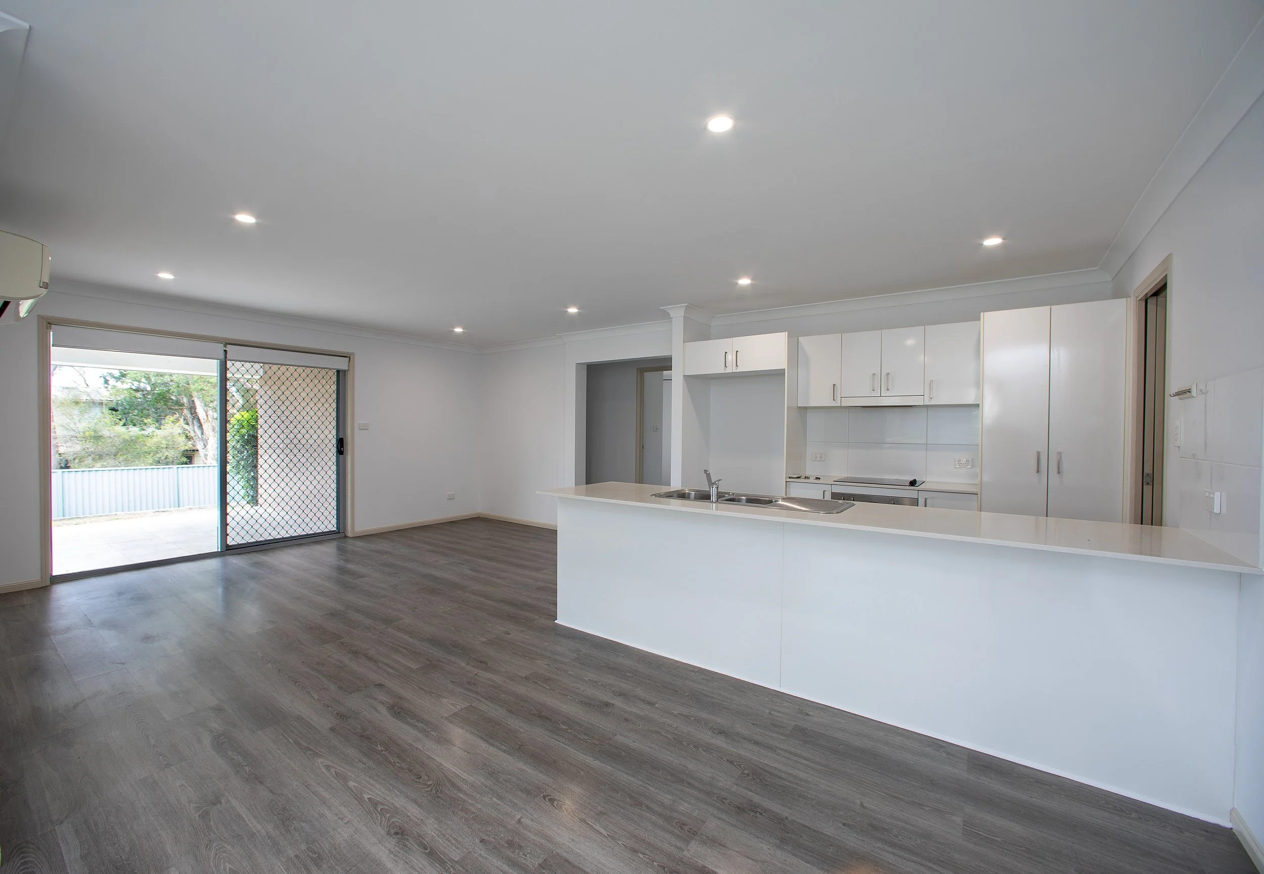 Empty modern open-plan living area and kitchen with white cabinets, grey flooring, and sliding glass door leading to outdoor space.