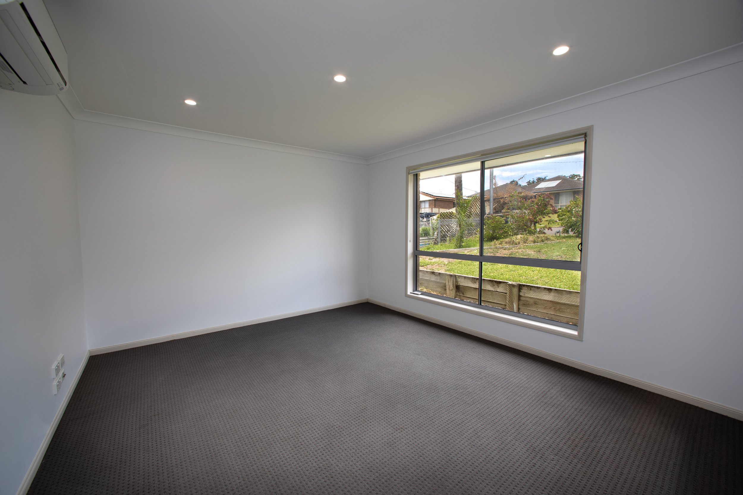 Empty room with gray carpet, white walls, large window showing a grassy yard and neighboring houses, ceiling lights, and an air conditioning unit.