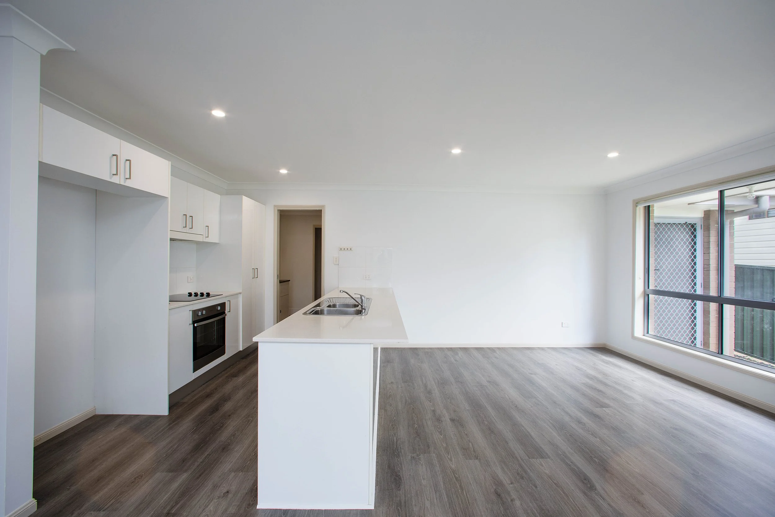 Empty living area with white kitchen counters and cabinets, hardwood floors, and large window letting in natural light.