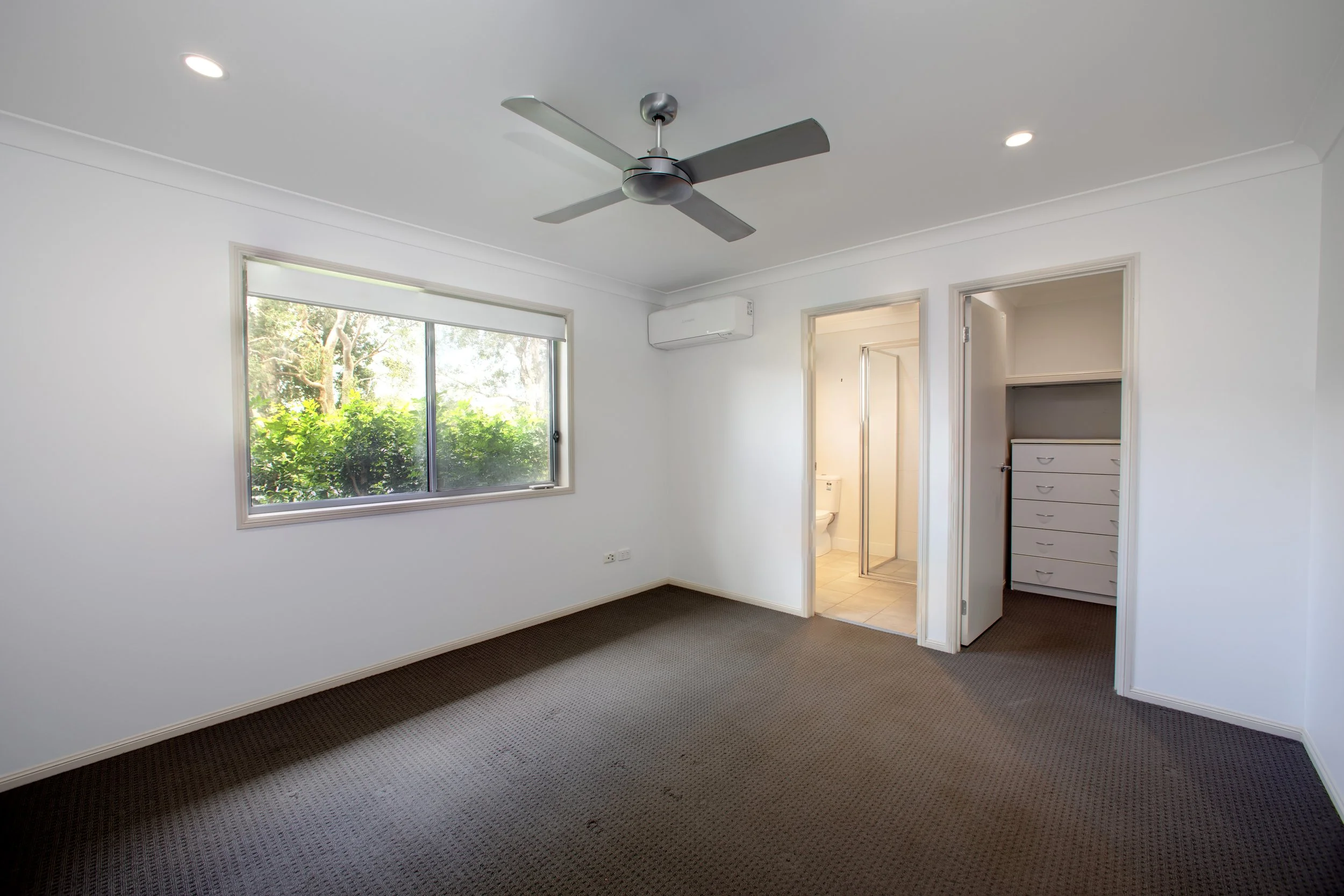 Empty bedroom with white walls, a large window with greenery outside, a ceiling fan, and an open closet with drawers.