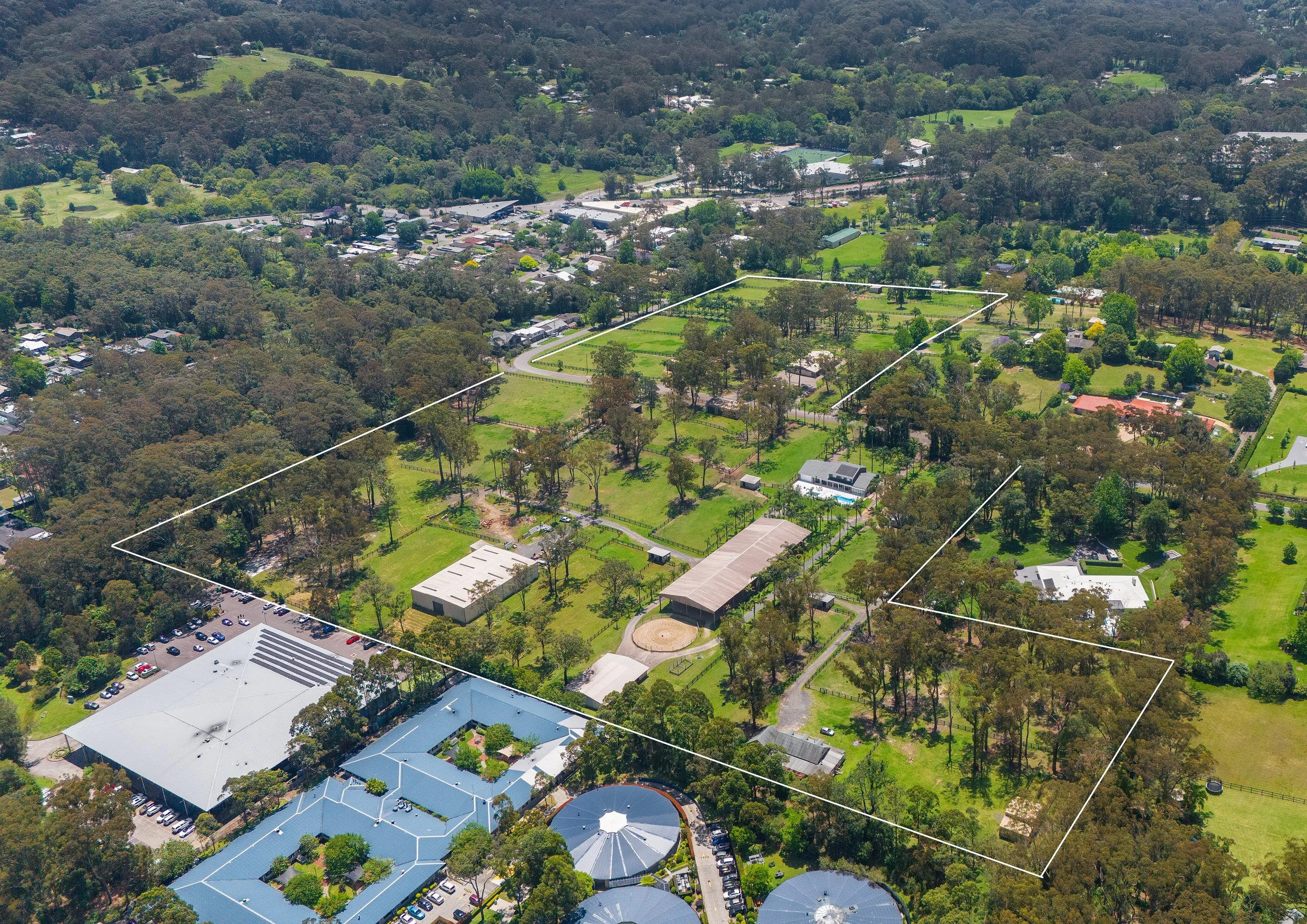 Aerial view of a large property outlined with white borders, showing green lawns, trees, several buildings, and parking areas surrounded by residential neighborhoods and wooded areas.