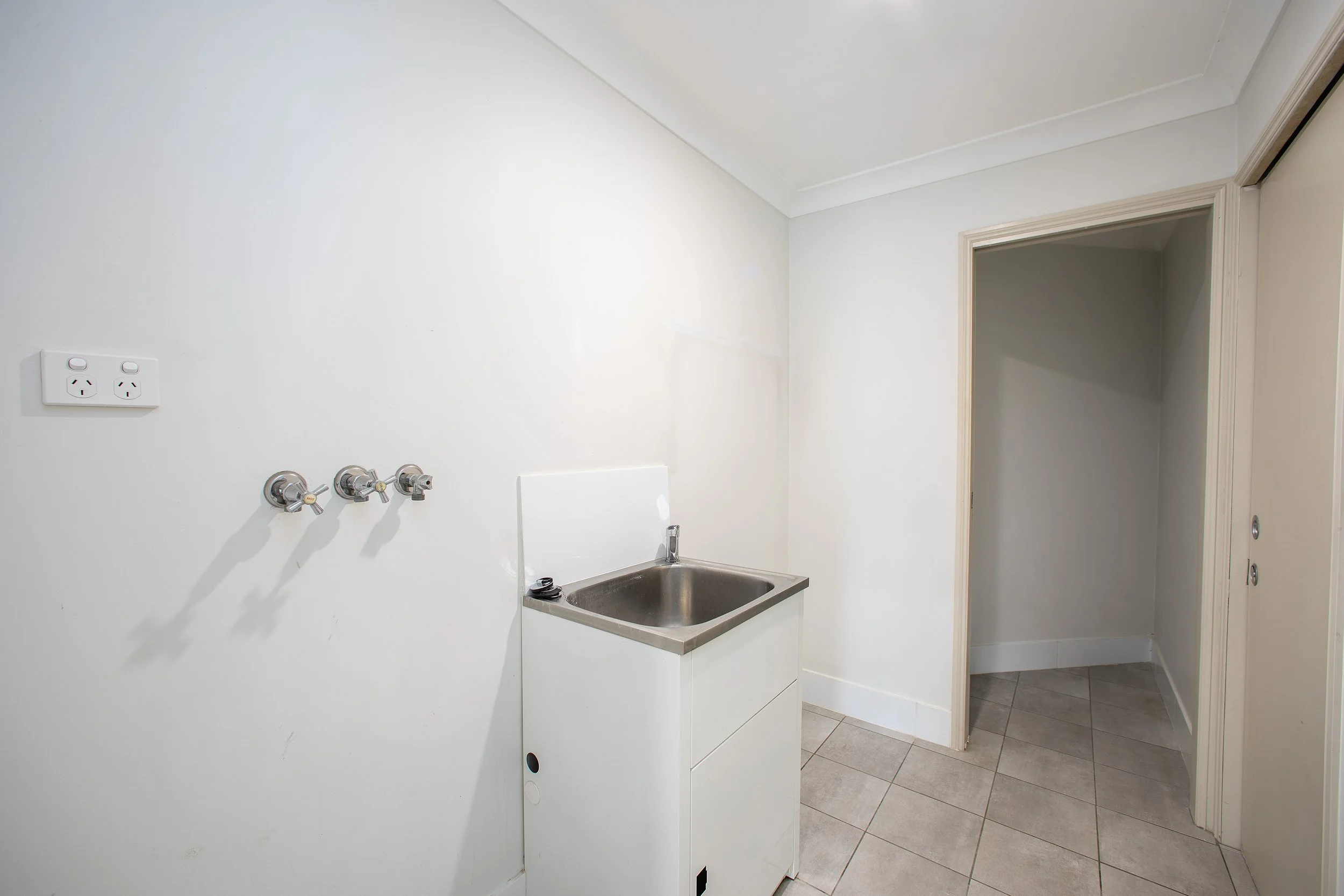 Empty laundry room with utility sink, wall power outlets, and hooks for hooks or supplies, beige tiled floor, plain white walls, and doorway leading to an empty closet or storage space.