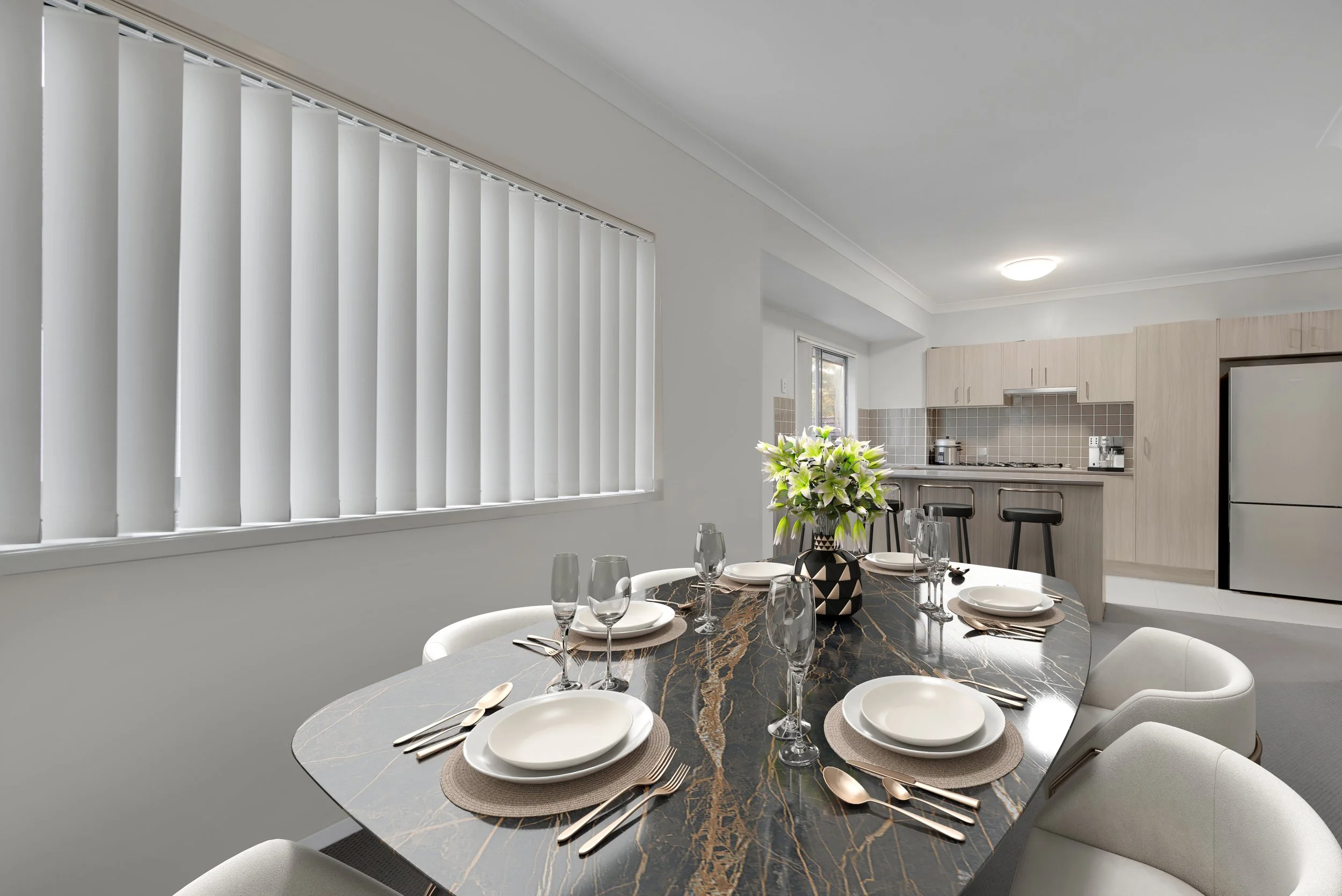 Dining table set with white plates, silverware, champagne glasses, and a black-and-white vase with white flowers, in a modern kitchen and dining area with white walls, vertical blinds, and a light wood kitchen. 