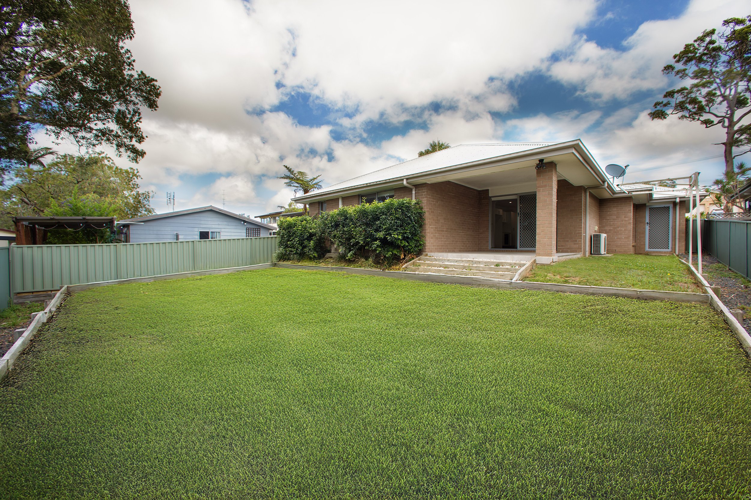 A backyard with a well-maintained green lawn, a brick house with a covered porch, bushes along the house, and trees on the sides under a partly cloudy sky.