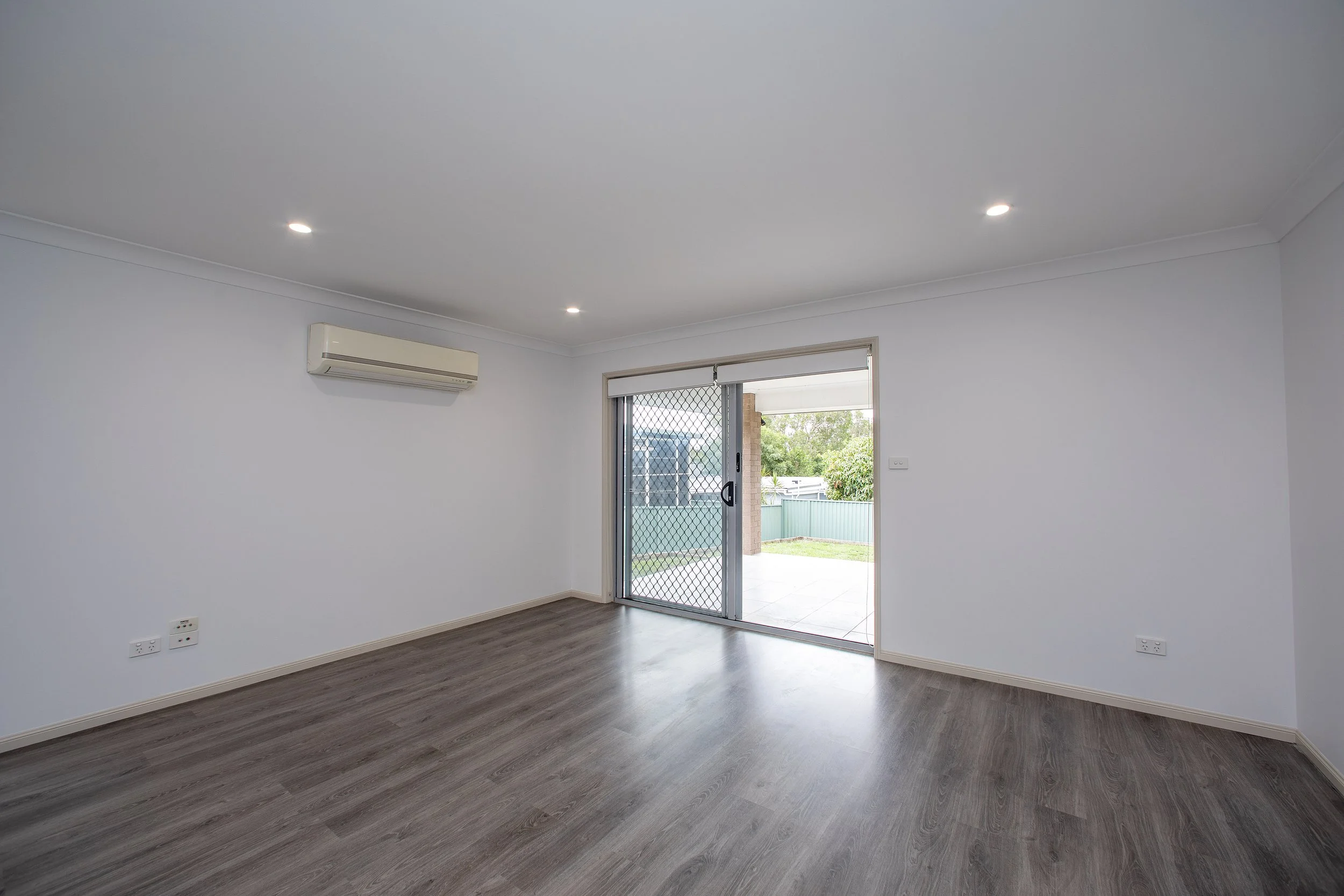 Empty living room with white walls, wood flooring, sliding glass door with security screen, view of backyard with grass and trees, ceiling lights, and wall-mounted air conditioning unit.