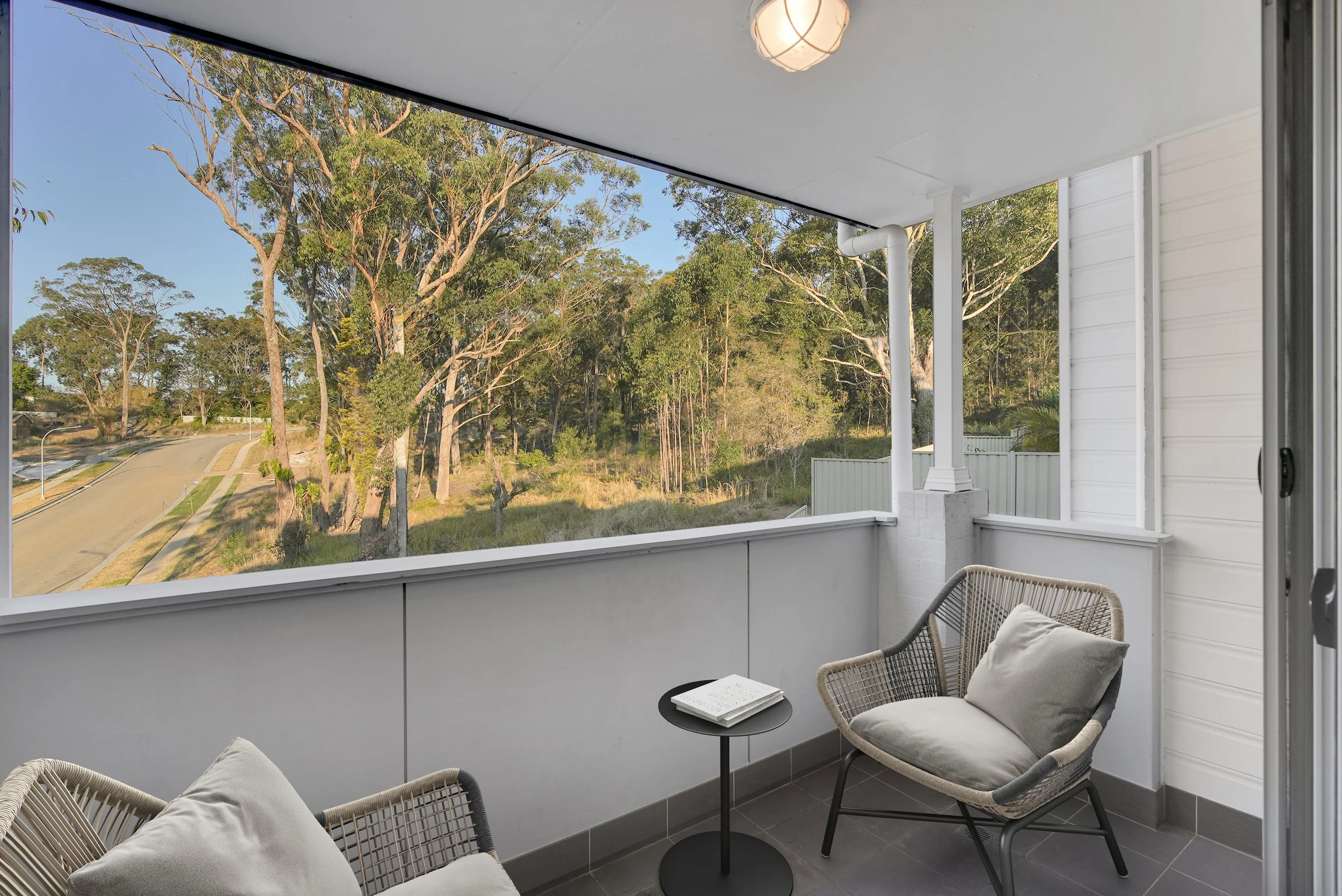 View from a balcony with two cushioned chairs, a small black side table holding a notebook, overlooking trees and a suburban street.