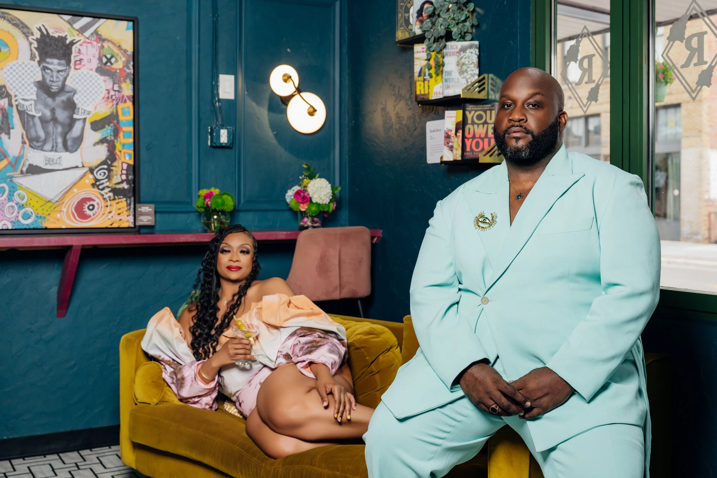 A woman with long braided hair is lying on a yellow velvet couch, holding a glass and looking at the camera. A man with a beard, dressed in a light blue suit, is standing nearby with his hands clasped, also looking at the camera. The background features a dark teal wall with modern art and a bookshelf with books, along with flowers and decorative objects.